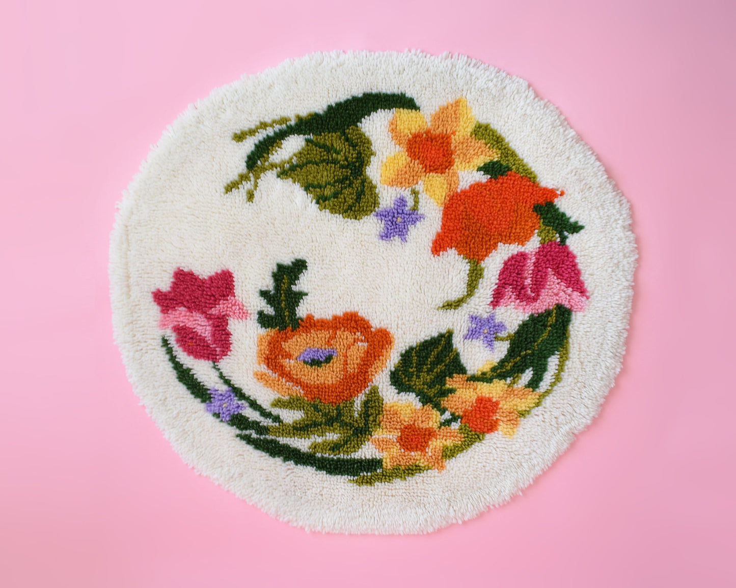 A vintage round floral latch hook rug that has cream white shaggy acrylic with a colorful floral motif in pinks, orange, yellow, purple and greens. The rug is on a pink background.