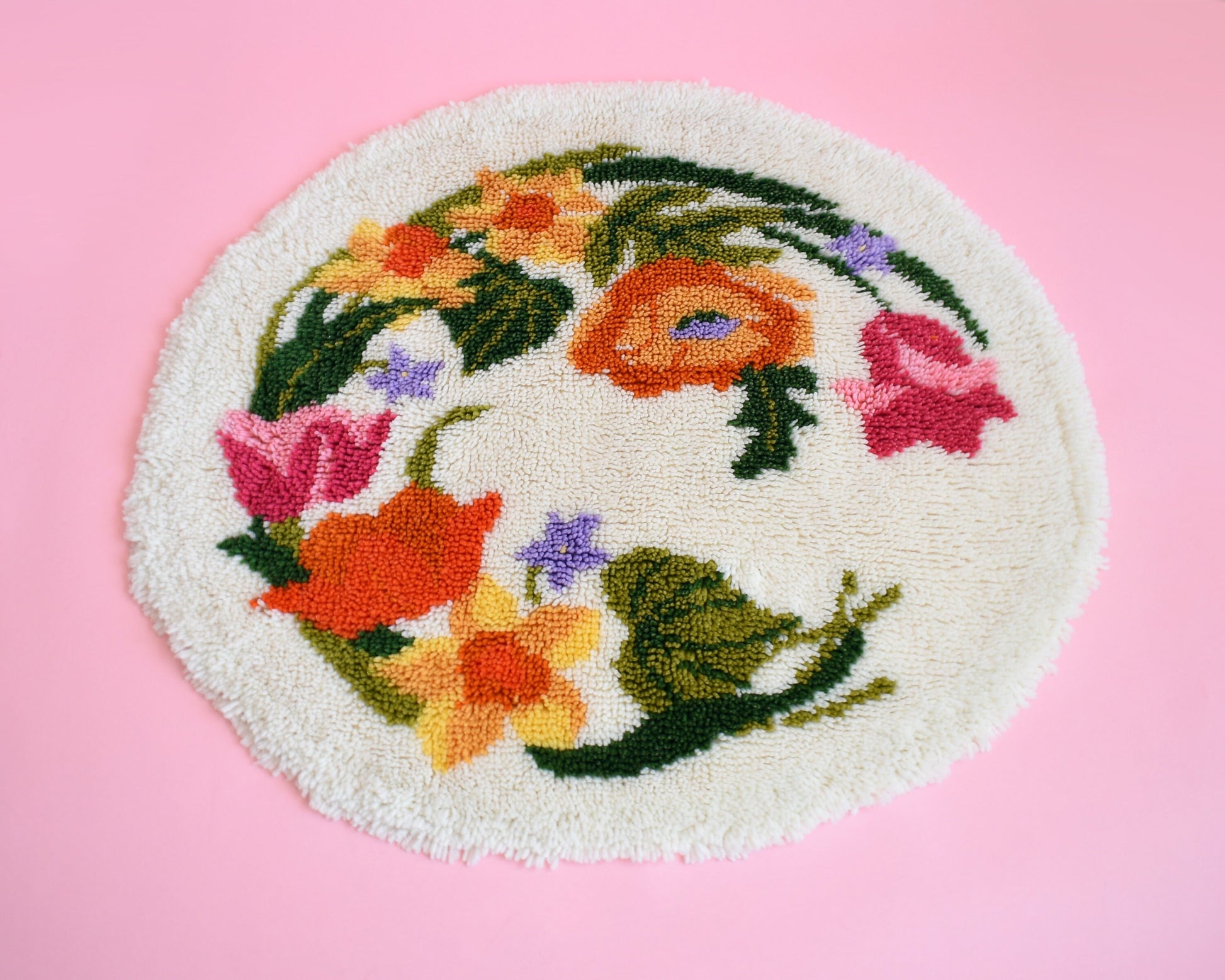 A vintage round floral latch hook rug that has cream white shaggy acrylic with a colorful floral motif in pinks, orange, yellow, purple and greens. The rug is on a pink background.