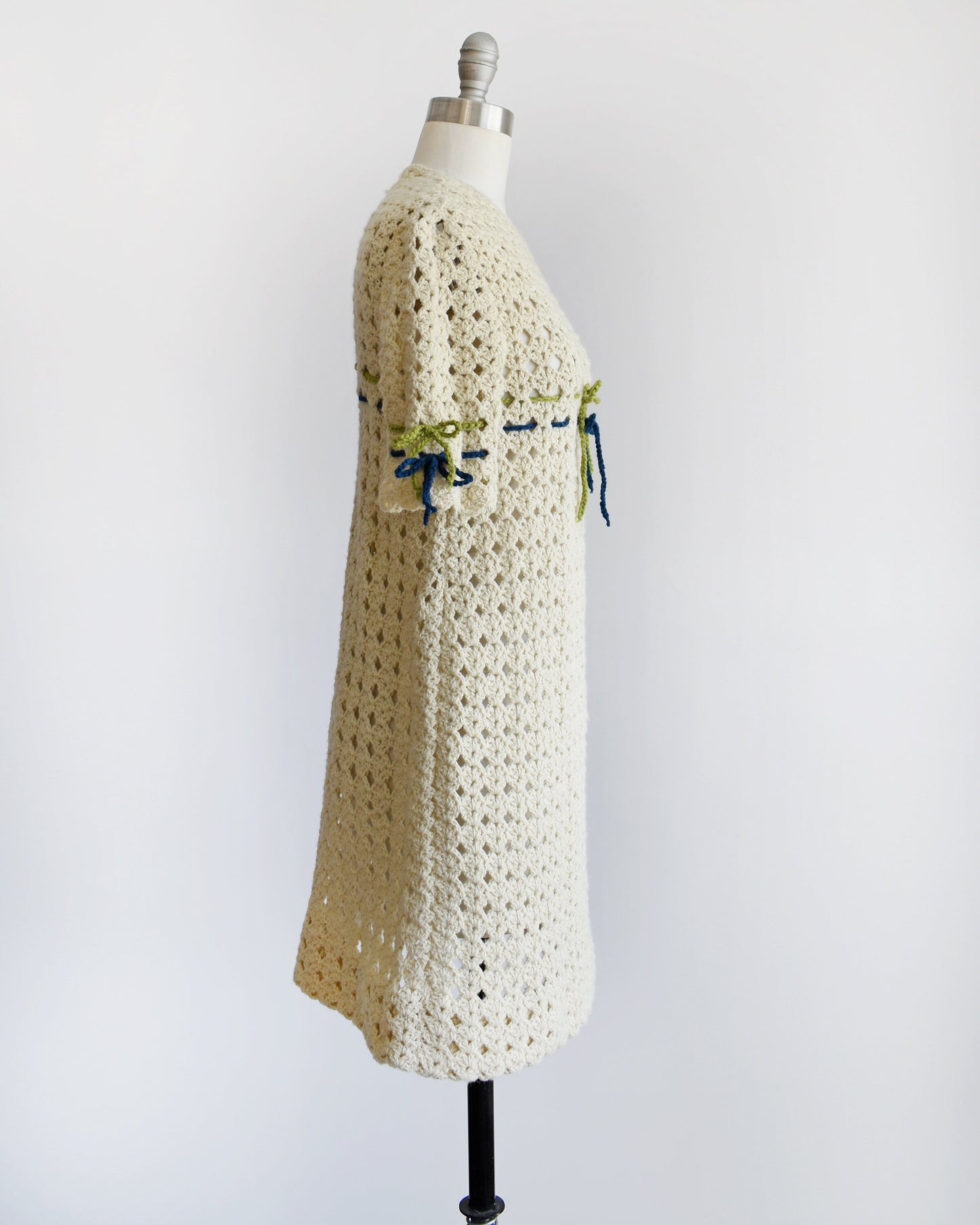 Side view of a vintage 70s crochet mini dress with a green and blue woven stripe on the front that ties into a bow, along with matching stripes and bows on the sleeves. The dress is on a dress form.