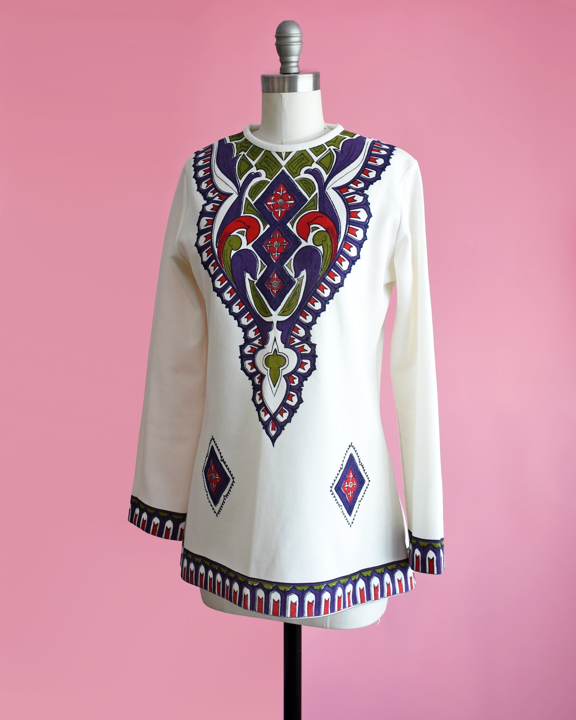 Side front view of a vintage 60s/70s white tunic with a purple, green, red, and black psychedelic print on the front. Matching print along the hem and long sleeve cuffs. The top is on a dress form.