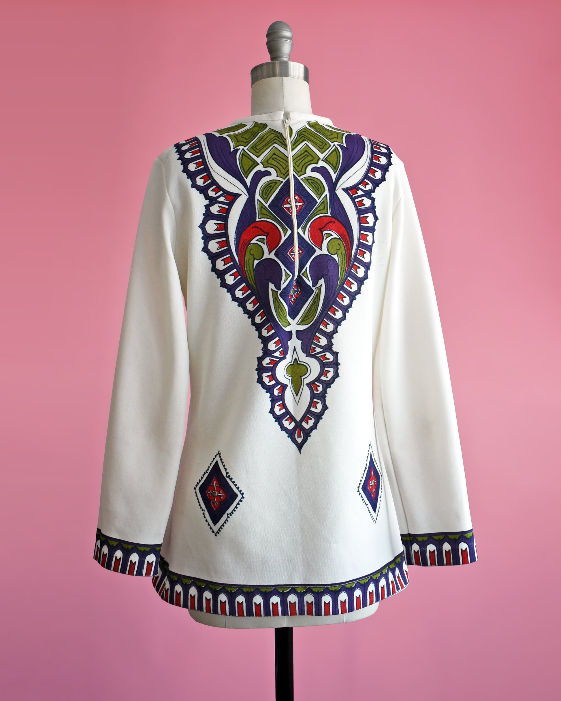 Back view of a vintage 60s/70s white tunic with a purple, green, red, and black psychedelic print on the back. Matching print along the hem and long sleeve cuffs. The top is on a dress form.