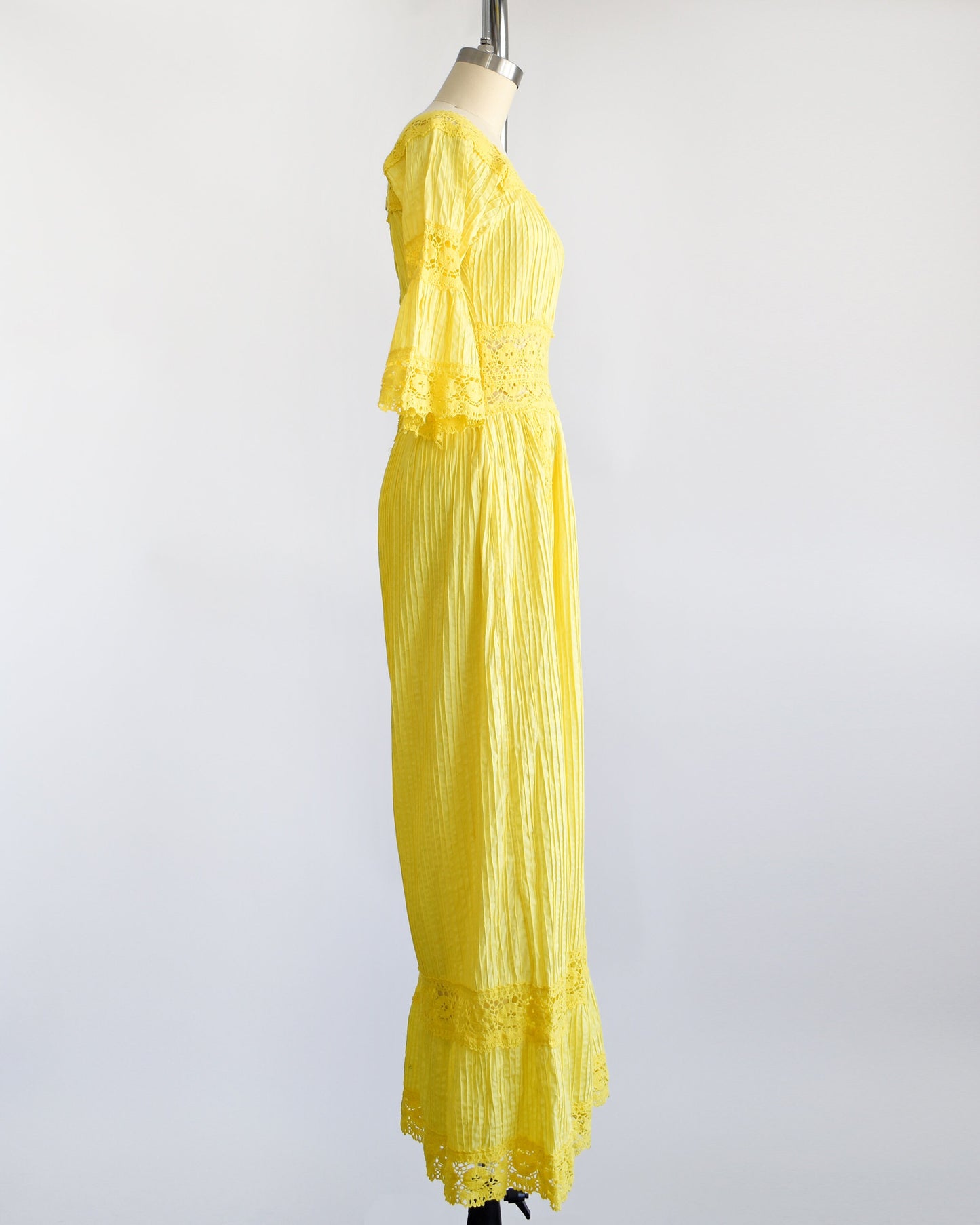 Side view of a vintage 70s yellow maxi dress with pin tuck pleats, along with lace trim around the collar, bell sleeves, waist, down the front, and around the hem. The dress is on a dress form.