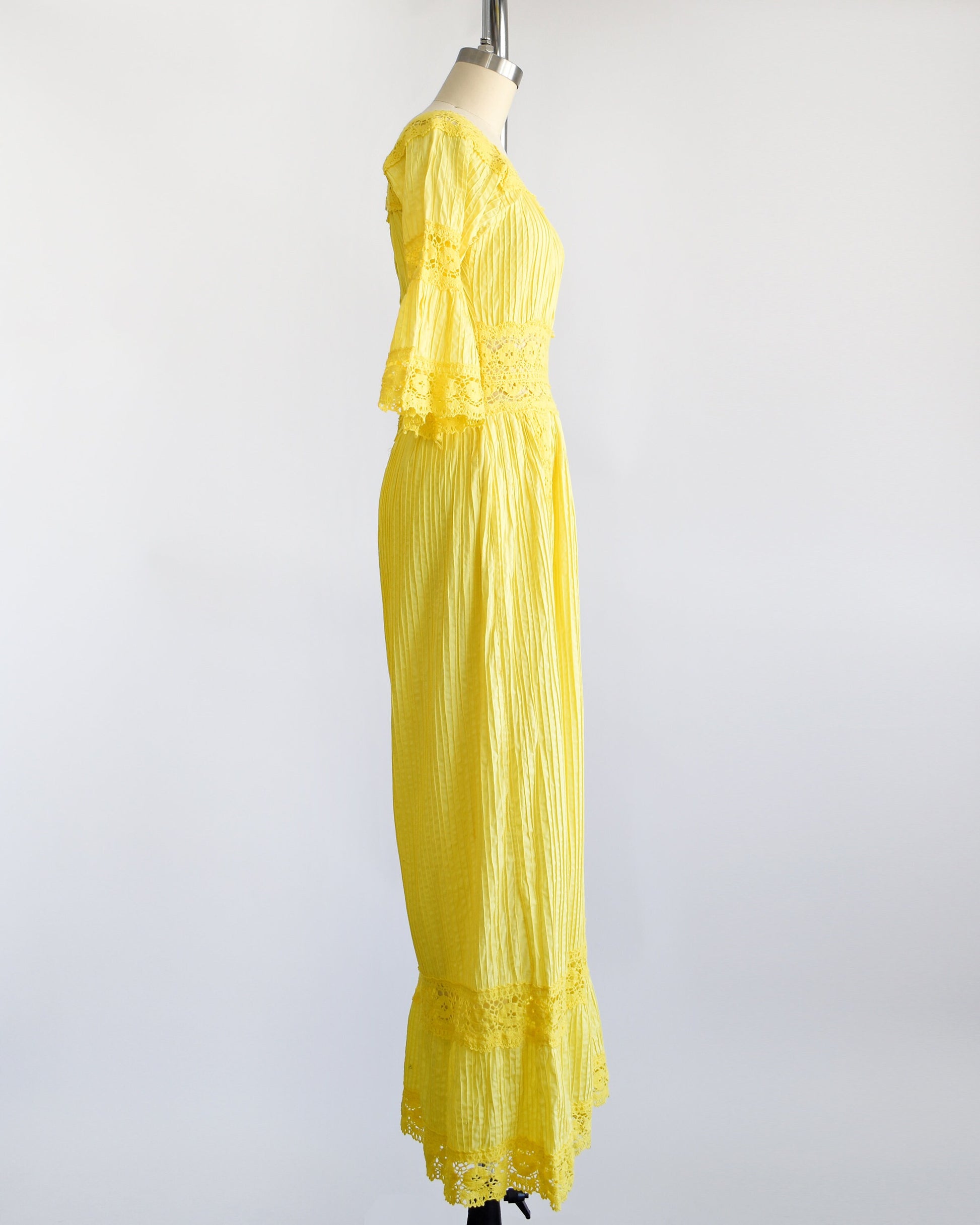 Side view of a vintage 70s yellow maxi dress with pin tuck pleats, along with lace trim around the collar, bell sleeves, waist, down the front, and around the hem. The dress is on a dress form.