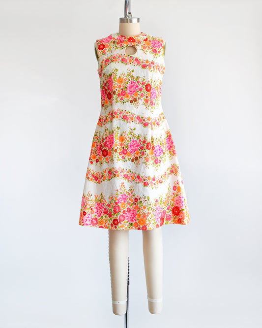 A vintage 60s/70s white dress that has a vibrant pink, orange, and green floral print in wavy horizontal stripes going down the dress. There is a circle cut out under the collar. The dress is on a dress form.