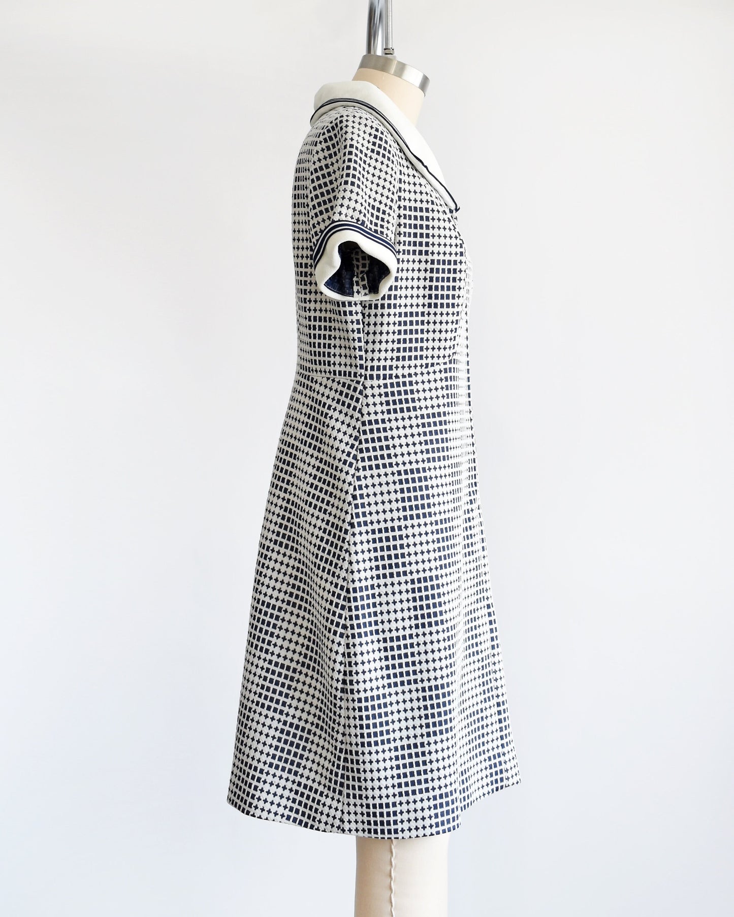 Side view of a vintage 70s mod dress that has a blue and white geometric print, white wide collar with navy trim, zip up front, and matching white and navy trim on the short sleeves. The dress is on a dress form.