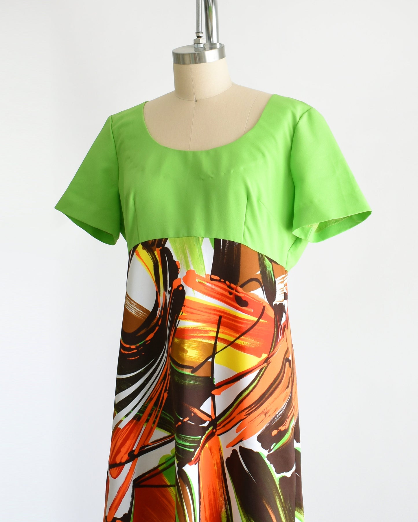 Side front view of a vintage 70s maxi dress with a lime green bodice with short sleeves and a maxi skirt that is white with an abstract browns, oranges, greens, and yellow large brushstroke print. The dress is on a dress form.