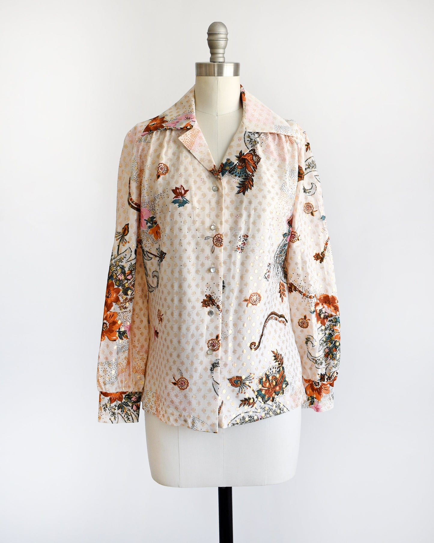 A vintage 70s light cream blouse that has a multicolored floral print and metallic gold connected polka dots all over. Pointy butterfly style collar. Pearly button up front. The blouse is on a dress form.