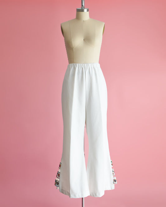 A pair of vintage 70s white bell bottom pants that have embroidered cut out detail on the side of the cuffs. The pants are on a dress form.