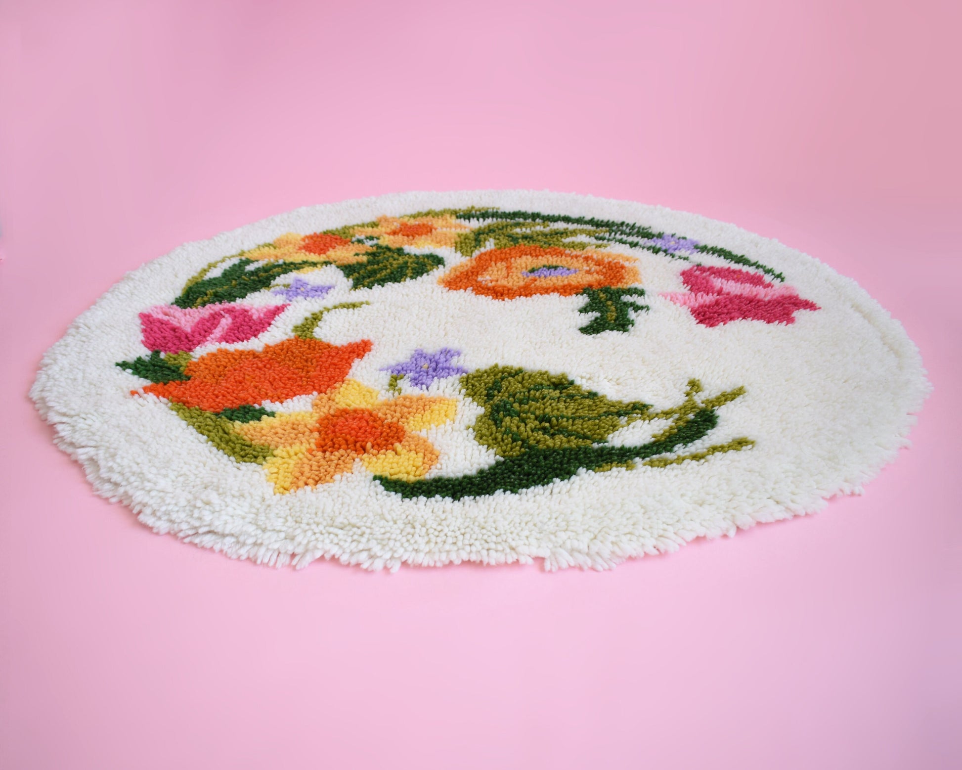 A view from the side of a vintage round floral latch hook rug that has cream white shaggy acrylic with a colorful floral motif in pinks, orange, yellow, purple and greens. The rug and the rug is on a pink background.