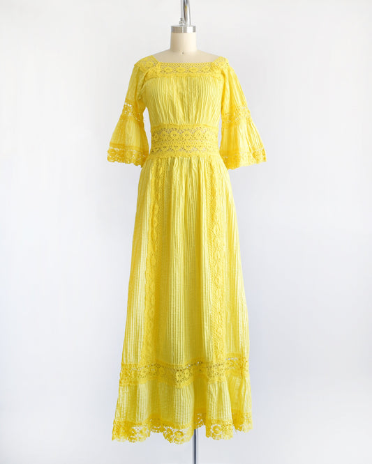 A vintage 70s yellow maxi dress with pin tuck pleats, along with lace trim around the collar, bell sleeves, waist, down the front, and around the hem. The dress is on a dress form.