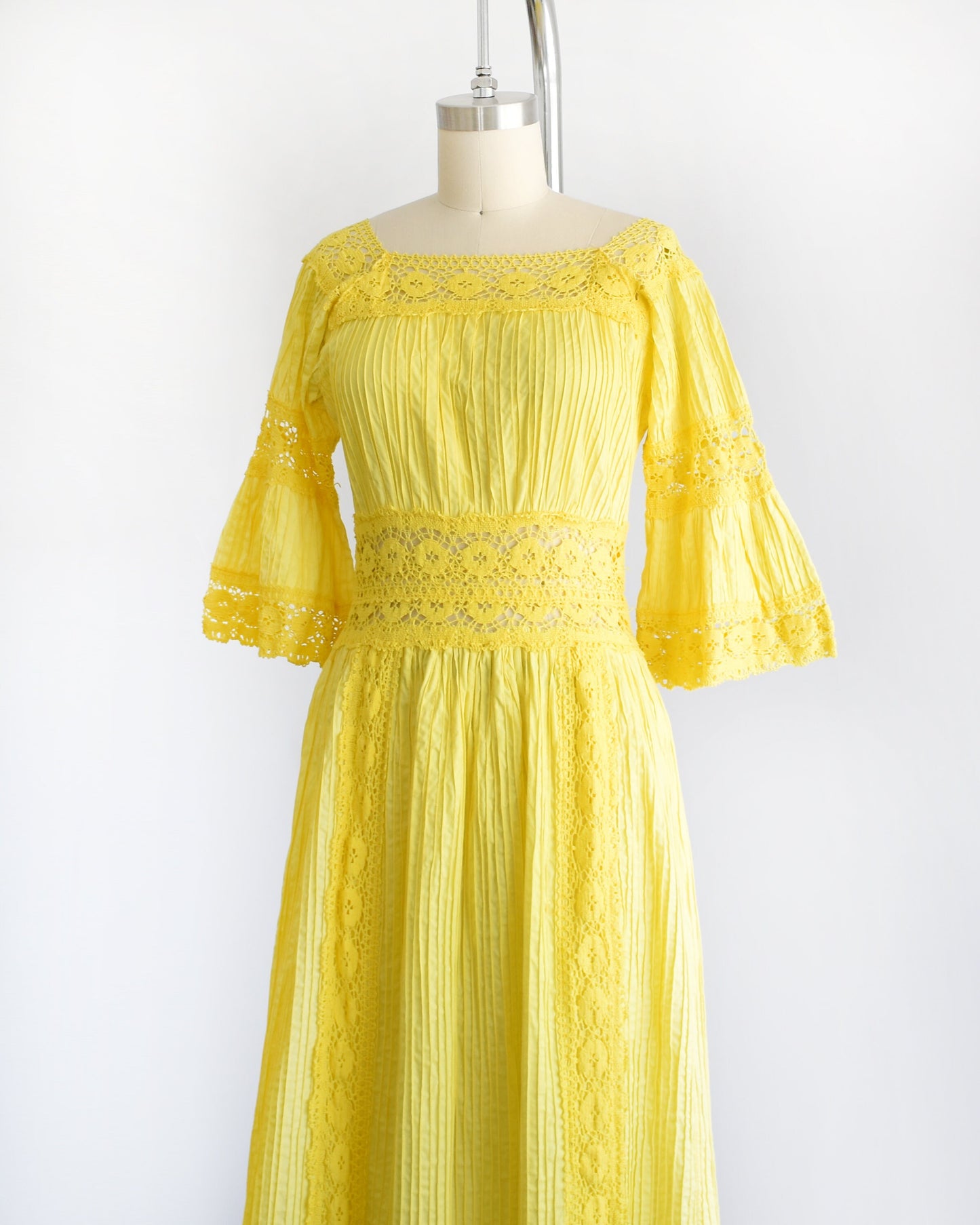 Side front view of a vintage 70s yellow maxi dress with pin tuck pleats, along with lace trim around the collar, bell sleeves, waist, down the front, and around the hem. The dress is on a dress form.