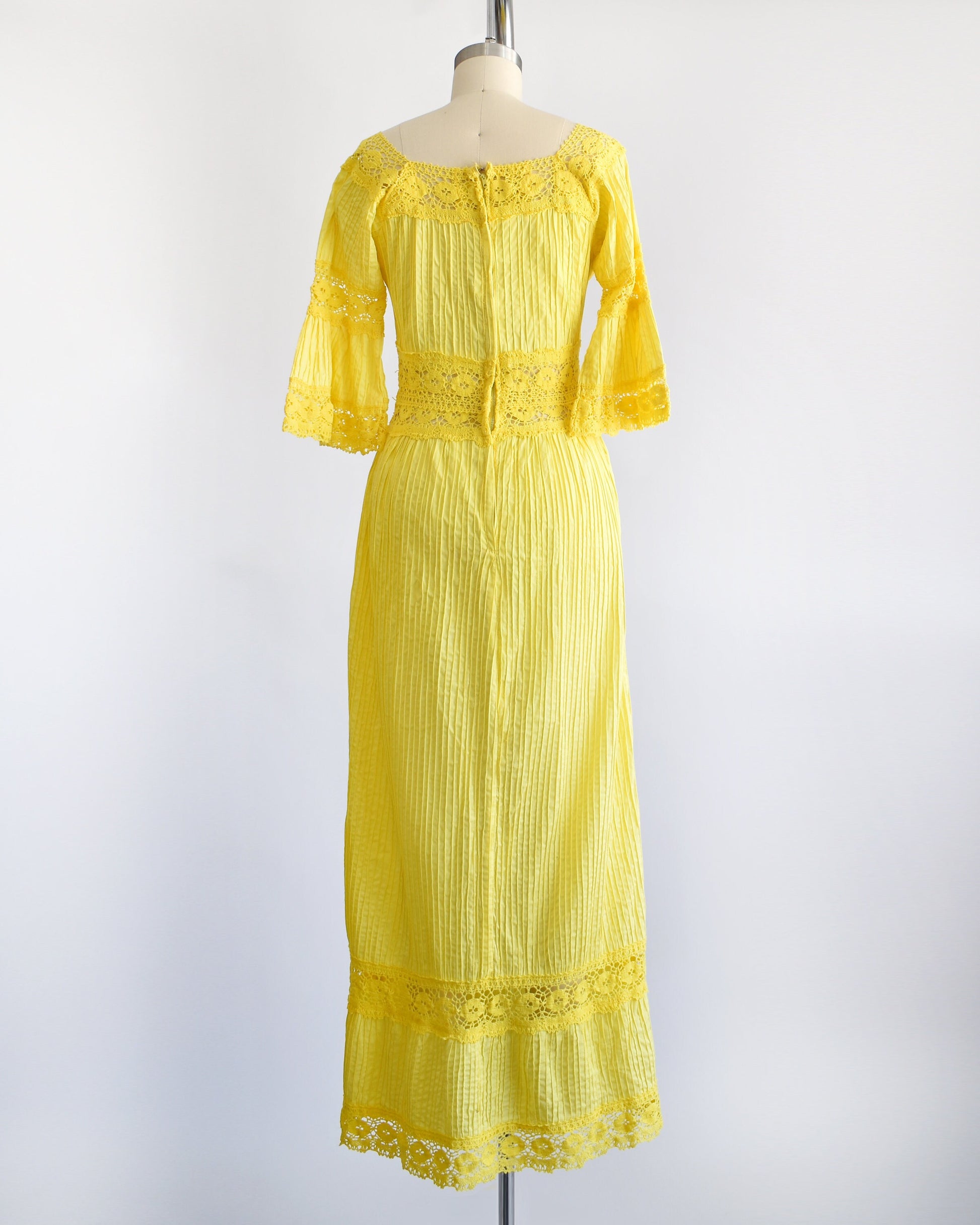 Back view of a vintage 70s yellow maxi dress with pin tuck pleats, along with lace trim around the collar, bell sleeves, waist, down the front, and around the hem. Zipper up the back. The dress is on a dress form.