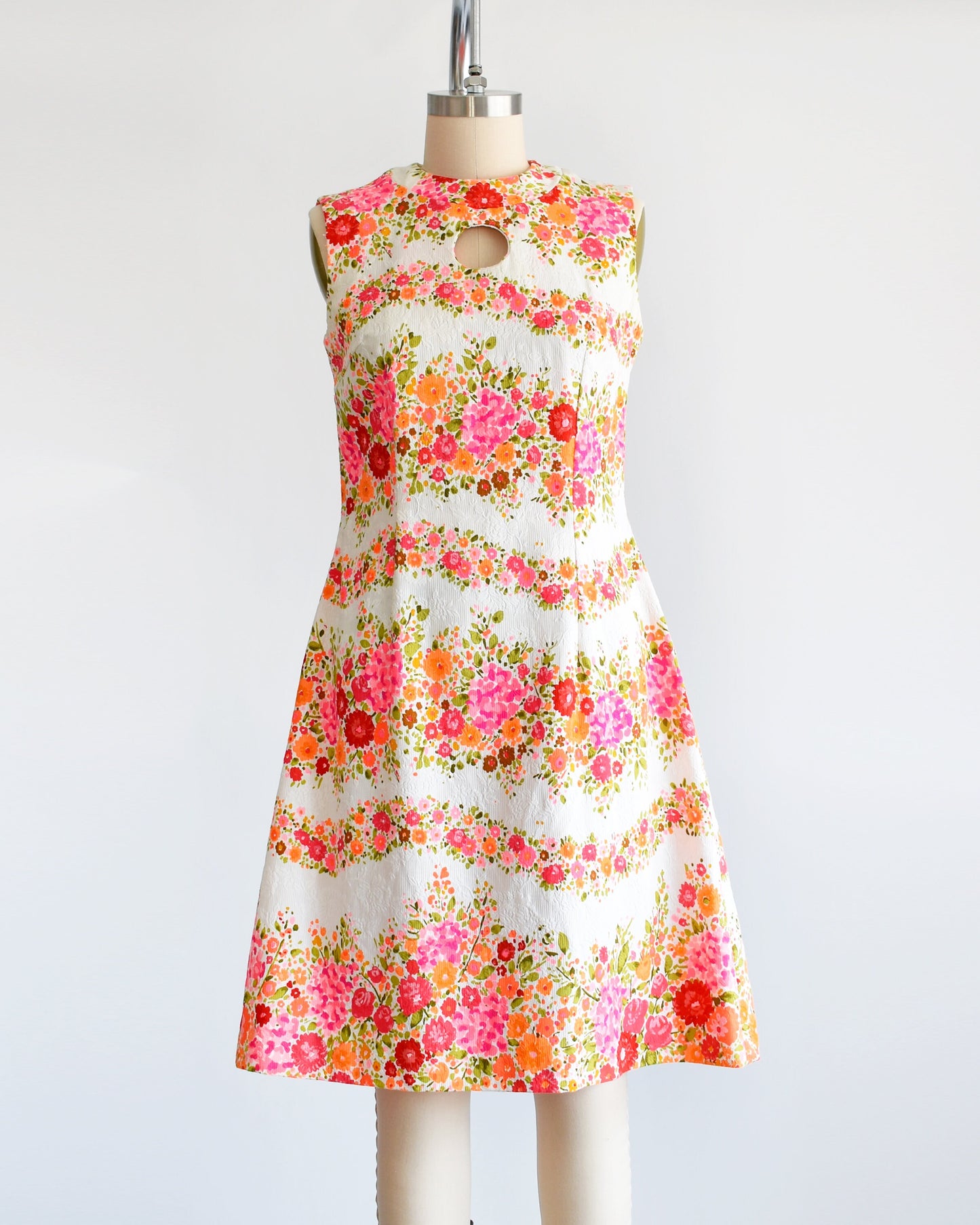 A vintage 60s/70s white dress that has a vibrant pink, orange, and green floral print in wavy horizontal stripes going down the dress. There is a circle cut out under the collar. The dress is on a dress form.