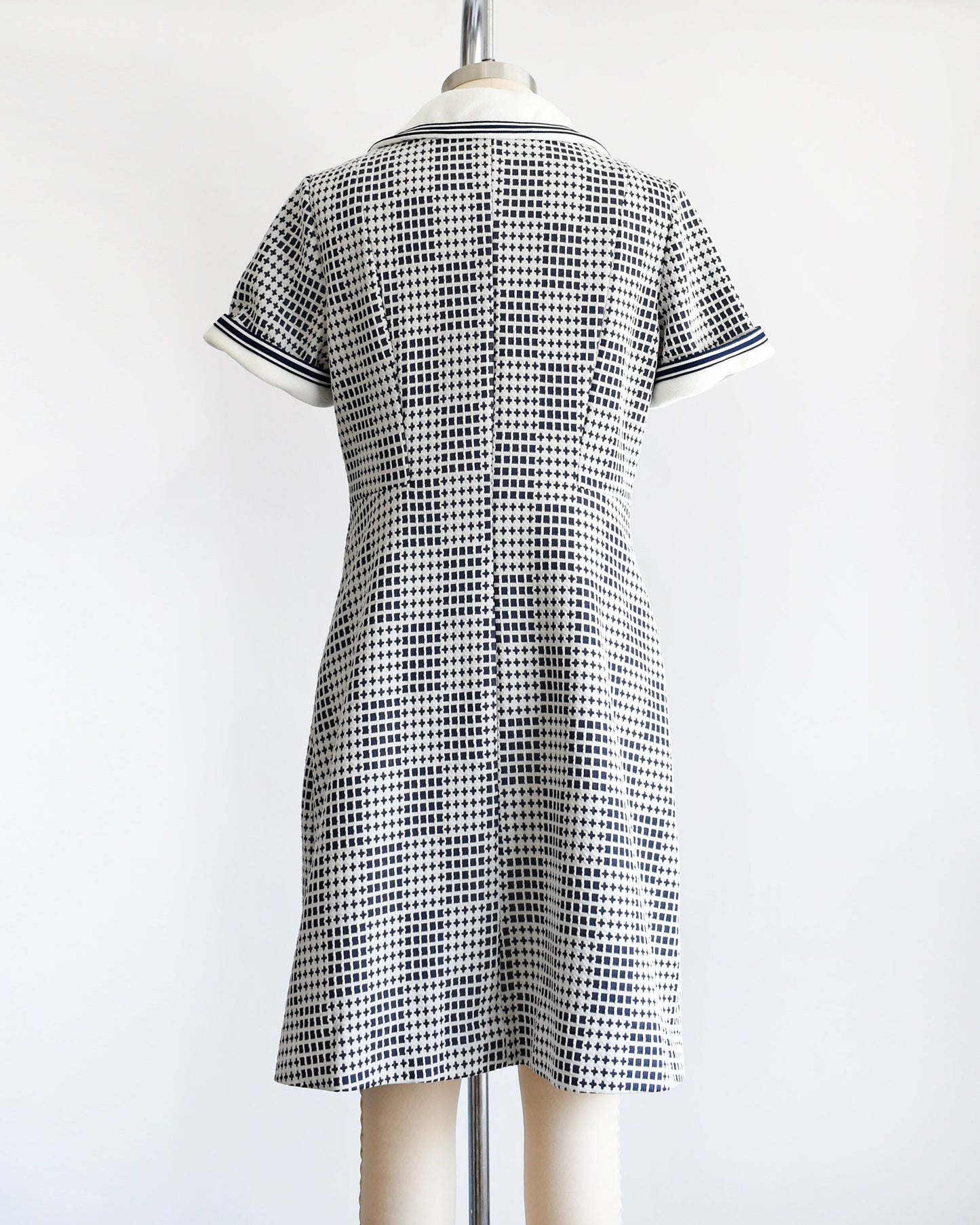 Back view of a A vintage 70s mod dress that has a blue and white geometric print, white wide collar with navy trim, and matching white and navy trim on the short sleeves. The dress is on a dress form.