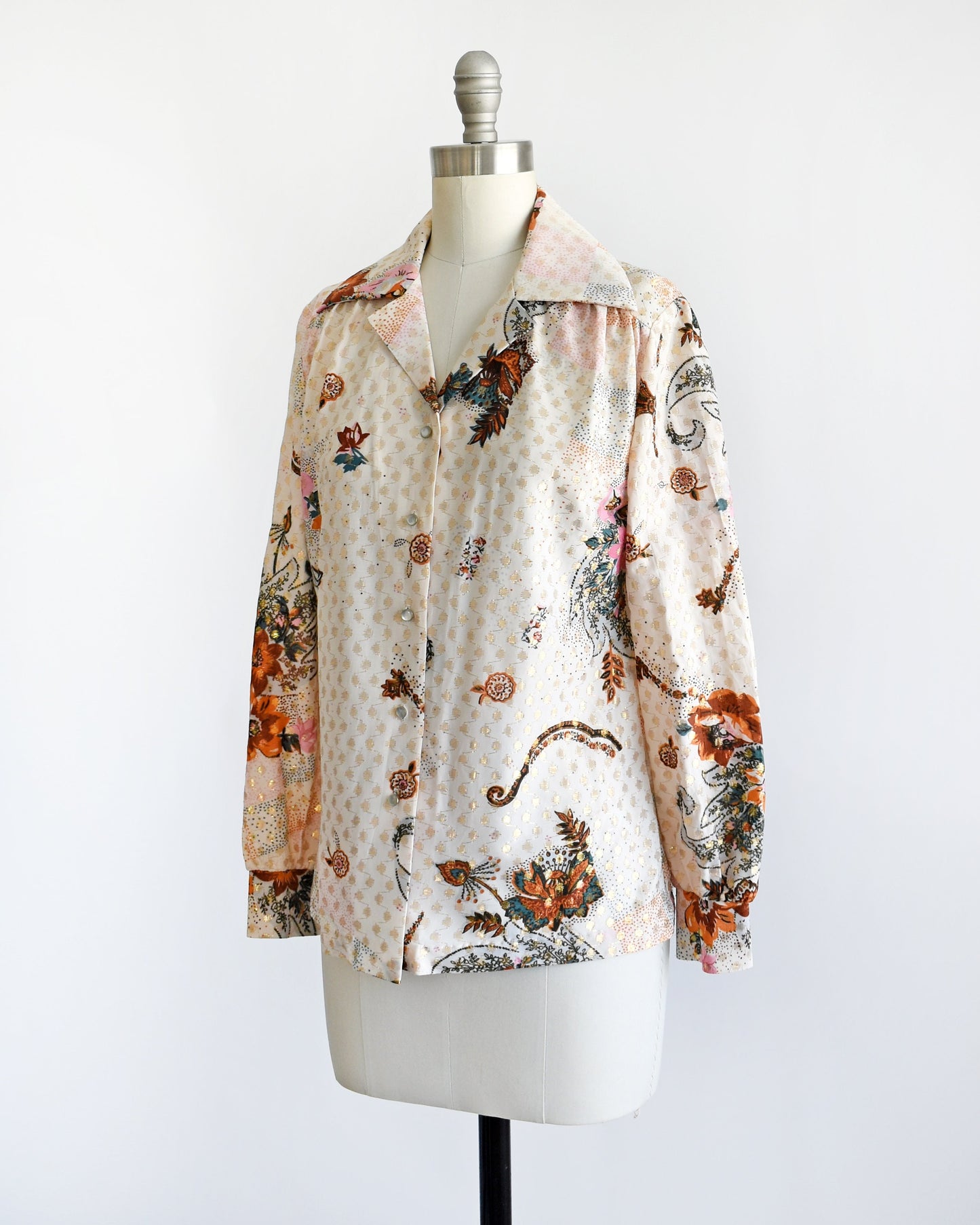 Side front view of a vintage 70s light cream blouse that has a multicolored floral print and metallic gold connected polka dots all over. Pointy butterfly style collar. Pearly button up front. The blouse is on a dress form.