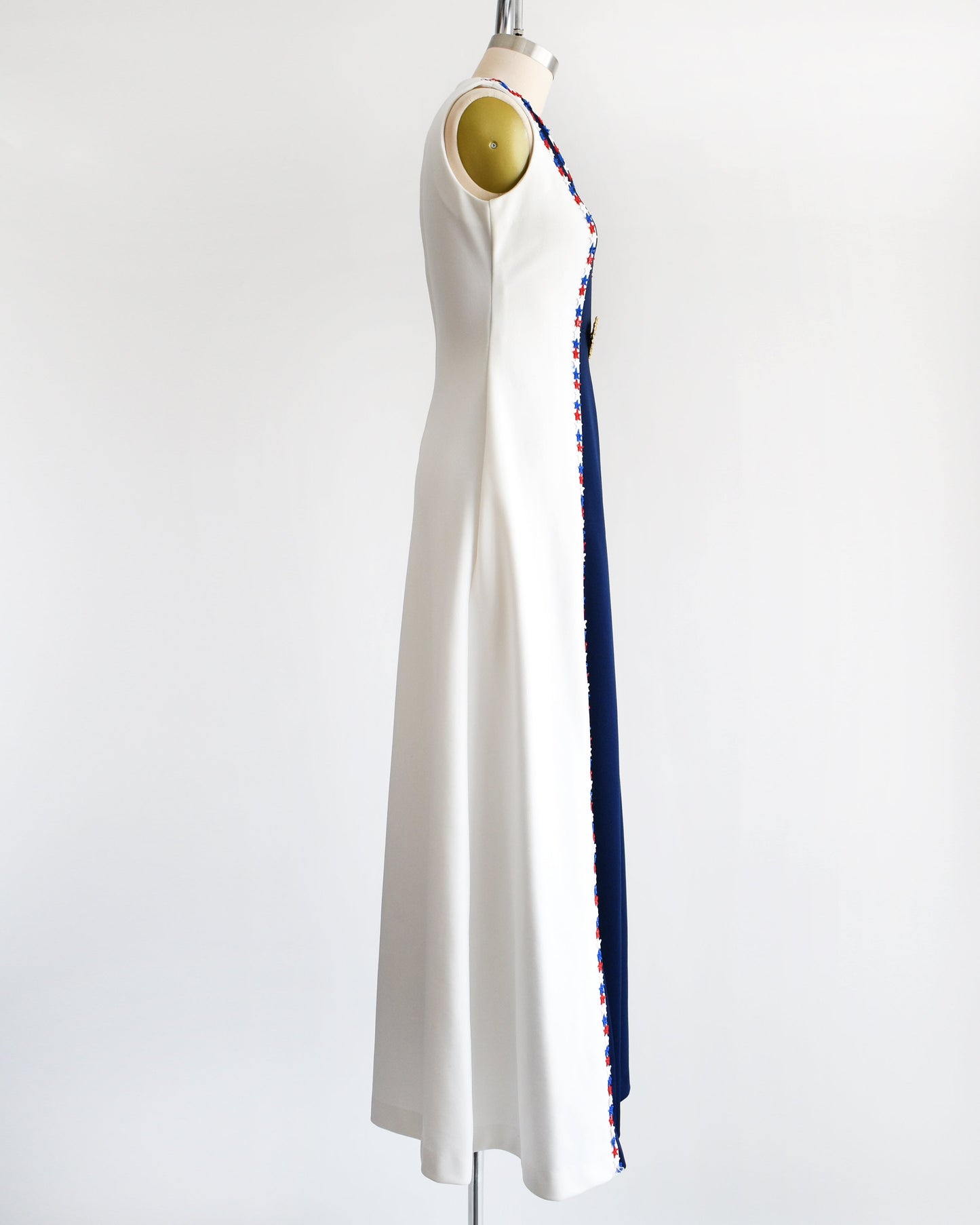 Side view of a vintage 70s maxi dress that is white with a long navy blue panel down the front. Embroidered stars applique around the collar and down the sides. A gold tone large brooch at the center of the dress.