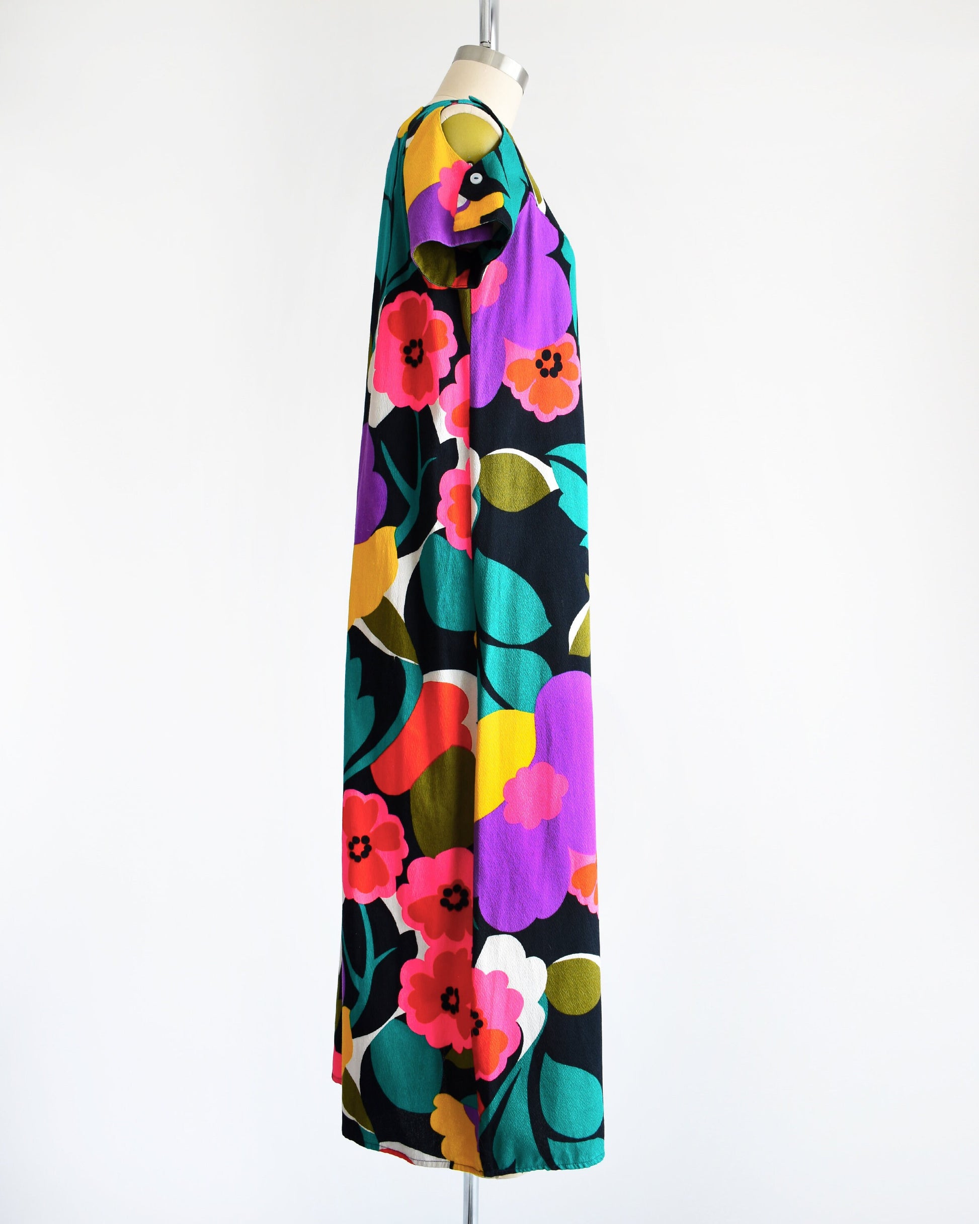 Side view of a vintage 70s Hawaiian floral maxi dress with black woven cotton that has a vibrant flower power print all over. Cold shoulder neckline with two buttons on each side, open shoulders, and short sleeves.