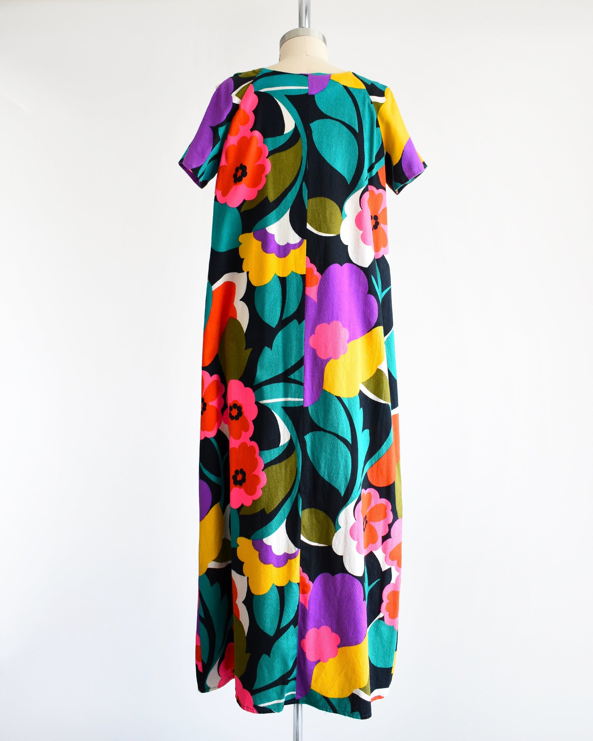 Back view of a vintage 70s Hawaiian floral maxi dress with black woven cotton that has a vibrant flower power print all over. Cold shoulder neckline with two buttons on each side, open shoulders, and short sleeves.