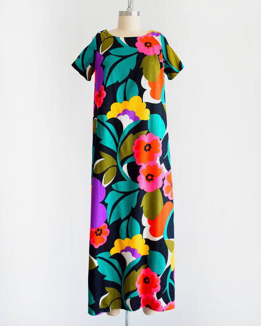 A vintage 70s Hawaiian floral maxi dress with black woven cotton that has a vibrant flower power print all over. Cold shoulder neckline with two buttons on each side, open shoulders, and short sleeves.