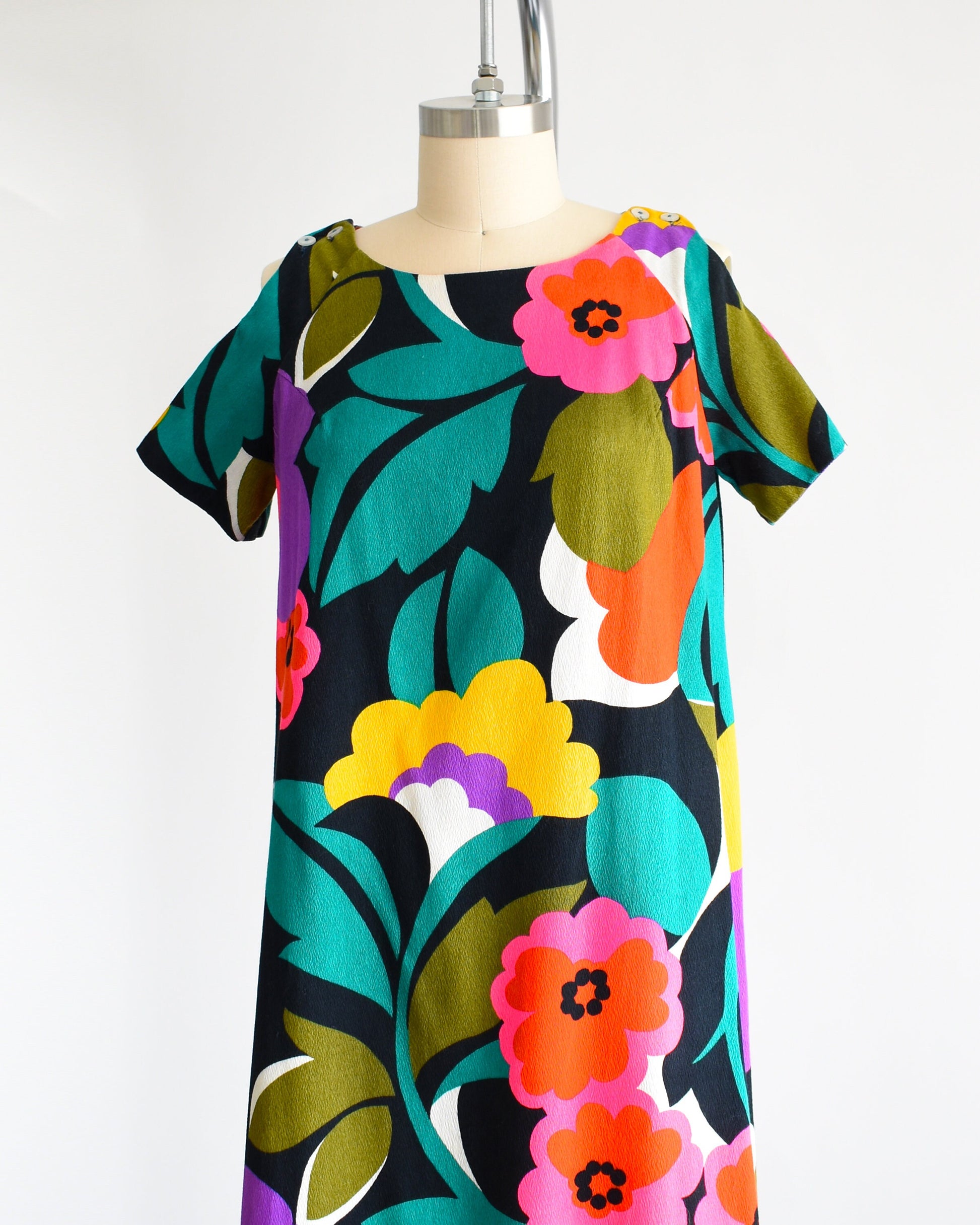 Side front view of a vintage 70s Hawaiian floral maxi dress with black woven cotton that has a vibrant flower power print all over. Cold shoulder neckline with two buttons on each side, open shoulders, and short sleeves.