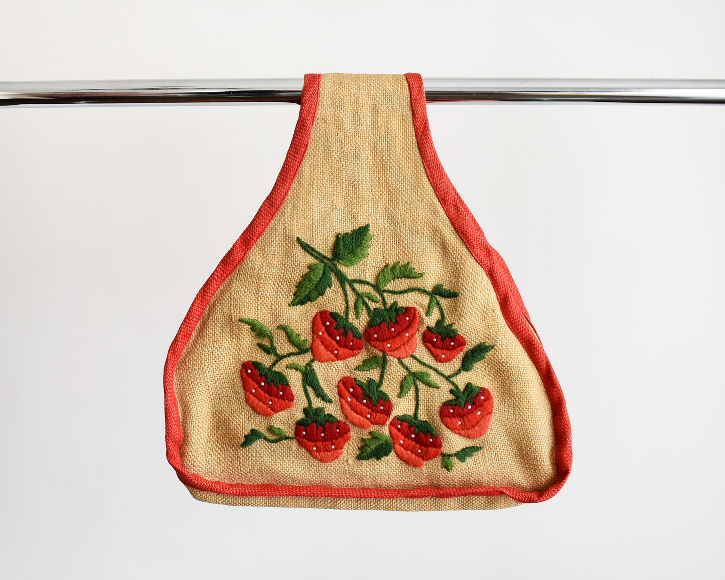 A vintage 60s burlap purse that has  embroidered strawberries on the front, with green leaves and small white pearl beaded seeds and red trim. The purse is being modeled on a pole
