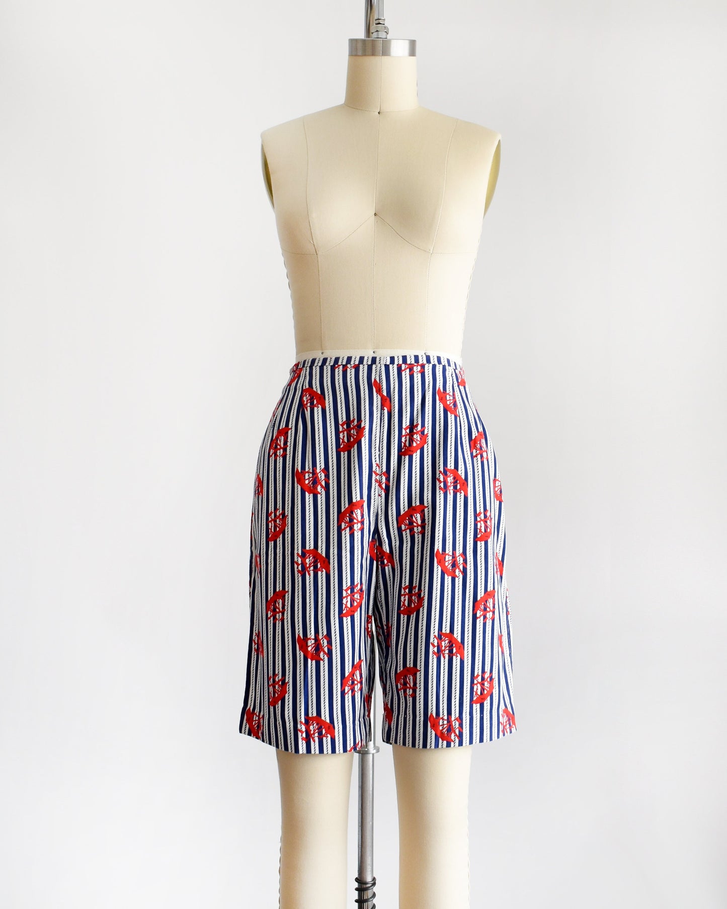 A pair of vintage 60s shorts that are navy blue and white vertical striped with a red clipper ship print. The shorts are modeled on a dress form. The shorts are cuffed in this photo.