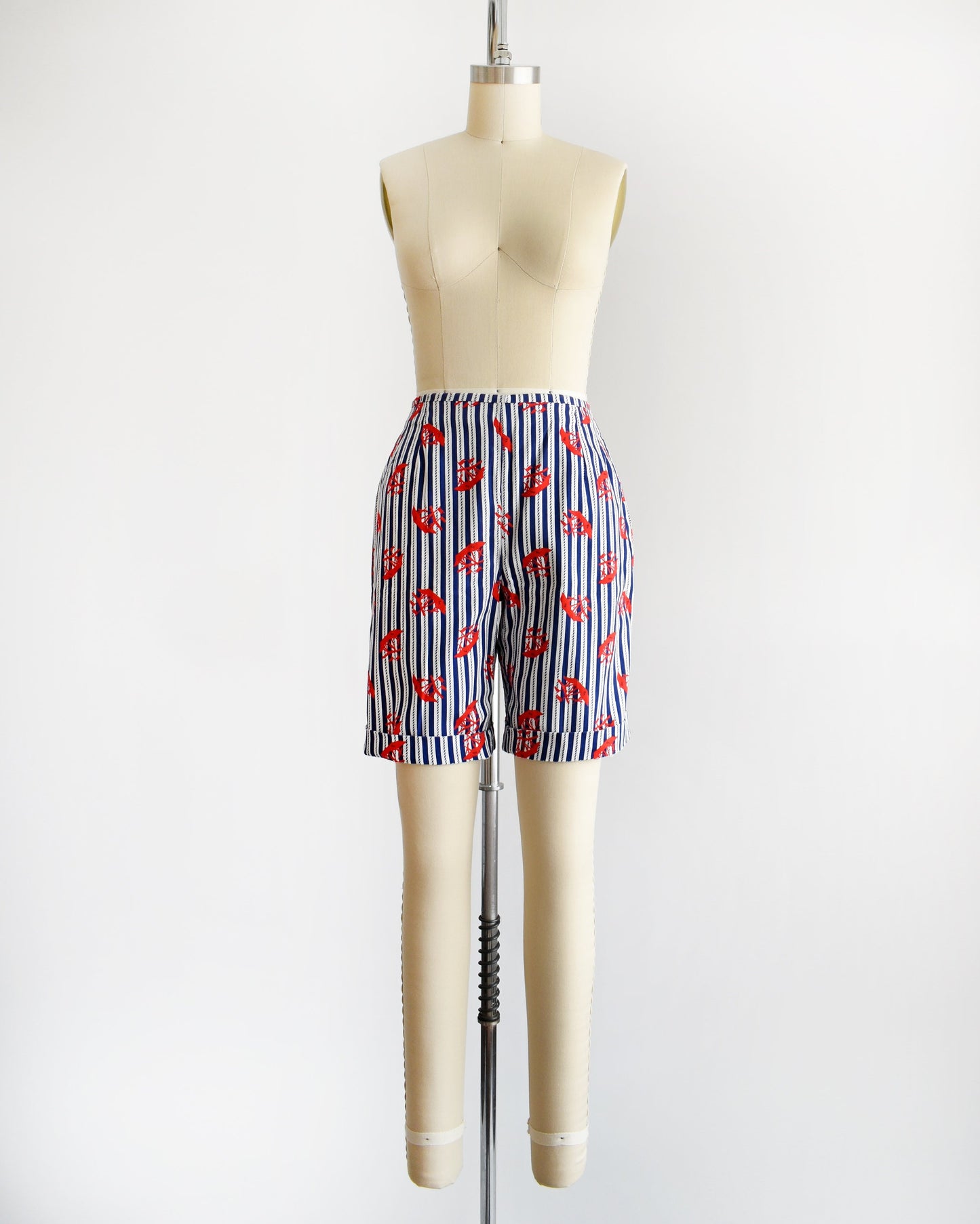 A pair of vintage 60s shorts that are navy blue and white vertical striped with a red clipper ship print. The shorts are modeled on a dress form. The shorts are cuffed in this photo.