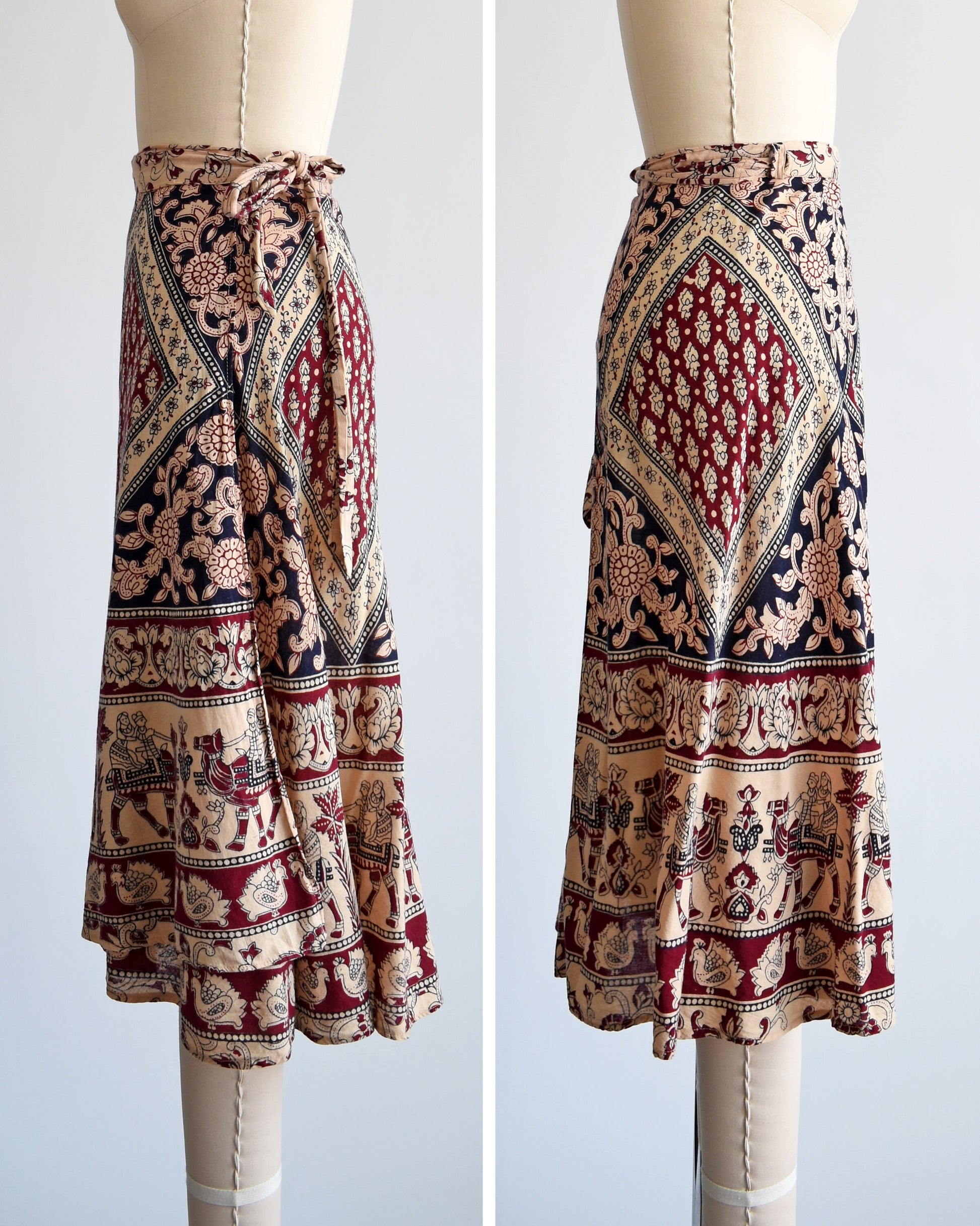 Side by side views of a vintage 1970s cotton wrap skirt that is beige with a large burgundy and black batik block floral print that covers the entire skirt, along border prints of people riding camels and a bird print. Skirt is on a dress form