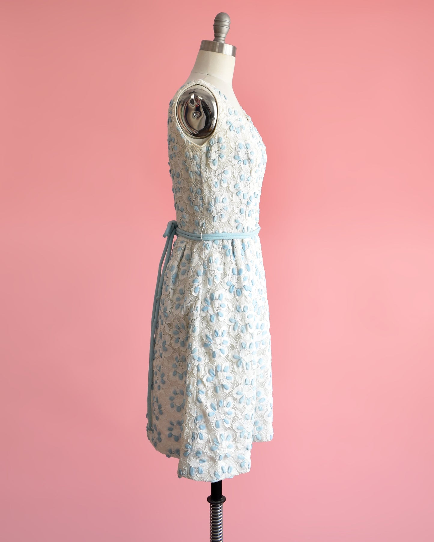 Side view of a vintage 60s white and blue floral lace dress that is sleeveless with a scoop neckline. Fitted waist with a matching double blue rope tie belt. Flared out skirt. Dress is modeled on a dress form