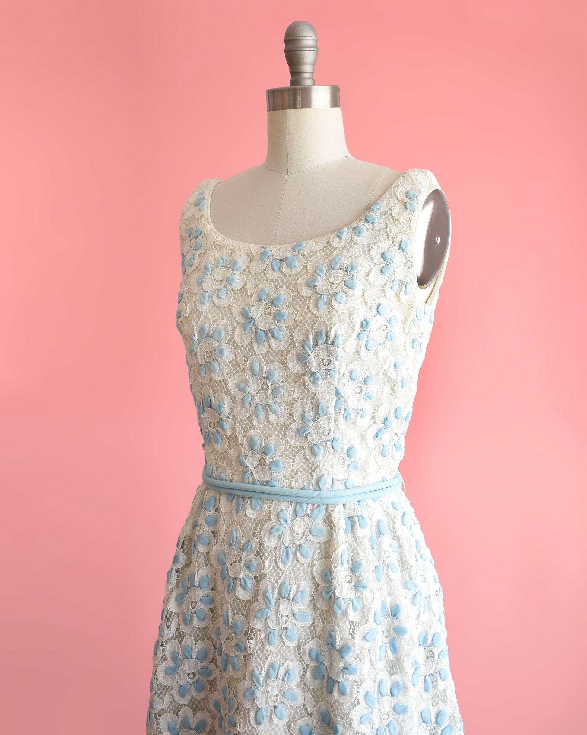 Side front view of a vintage 60s white and blue floral lace dress that is sleeveless with a scoop neckline. Fitted waist with a matching double blue rope tie belt. Flared out skirt. Dress is modeled on a dress form