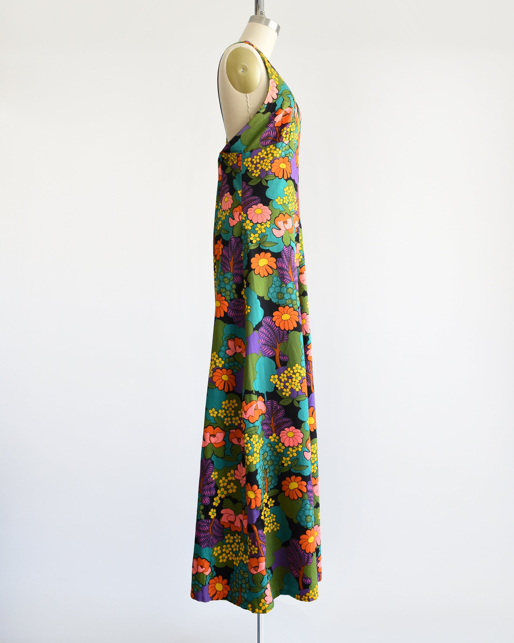 Side view of a vintage 70s floral maxi dress that is black and has a vibrant flower power print in greens, blue, peach, orange, yellow, purple, and white. V-neckline. Dress is on dress form.
