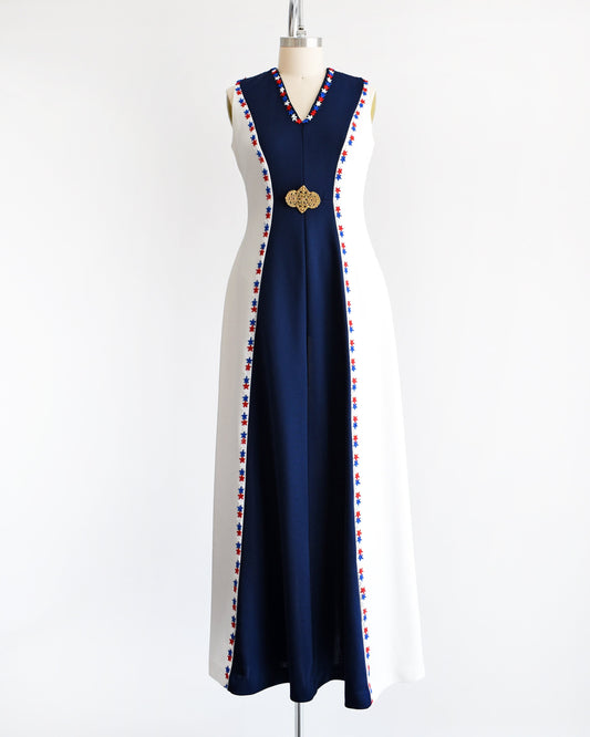 A vintage 70s maxi dress that is white with a long navy blue panel down the front. V-neckline with red, white, and blue embroidered stars applique around the collar and down the sides. A gold tone large brooch at the center of the dress.