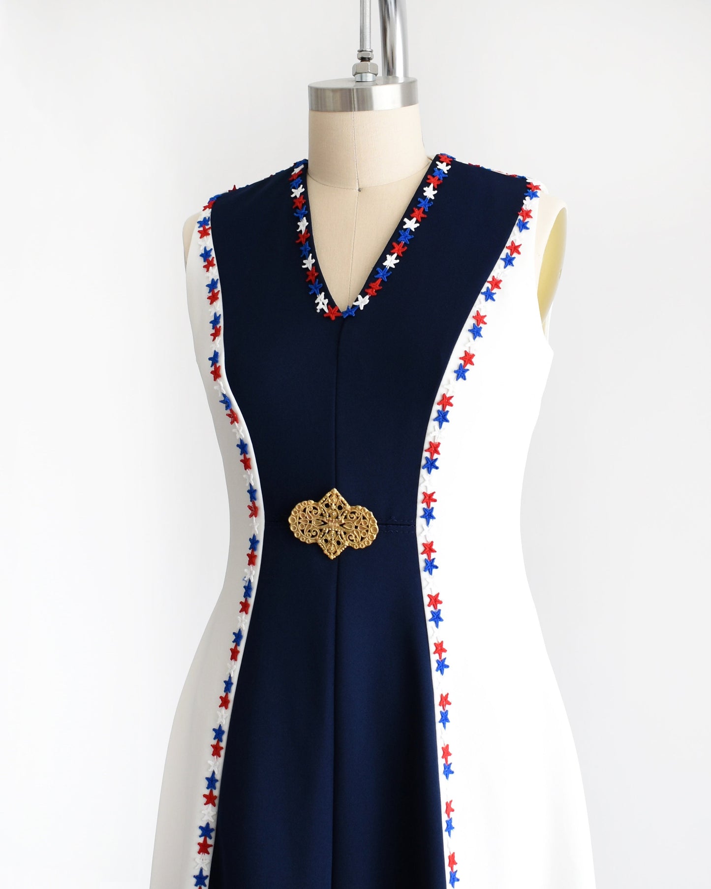 Side front view of a vintage 70s maxi dress that is white with a long navy blue panel down the front. V-neckline with red, white, and blue embroidered stars applique around the collar and down the sides. A gold tone large brooch at the waist