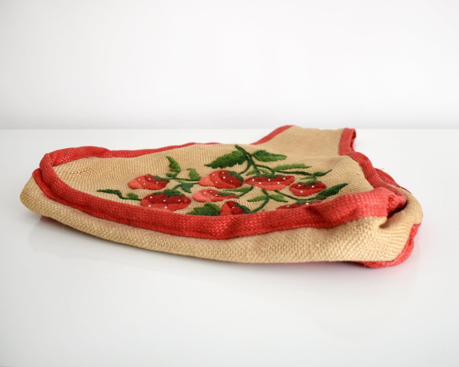 Side view of a vintage 60s burlap purse that has  embroidered strawberries on the front, with green leaves and small white pearl beaded seeds and red trim. The purse is laying flat on a white table