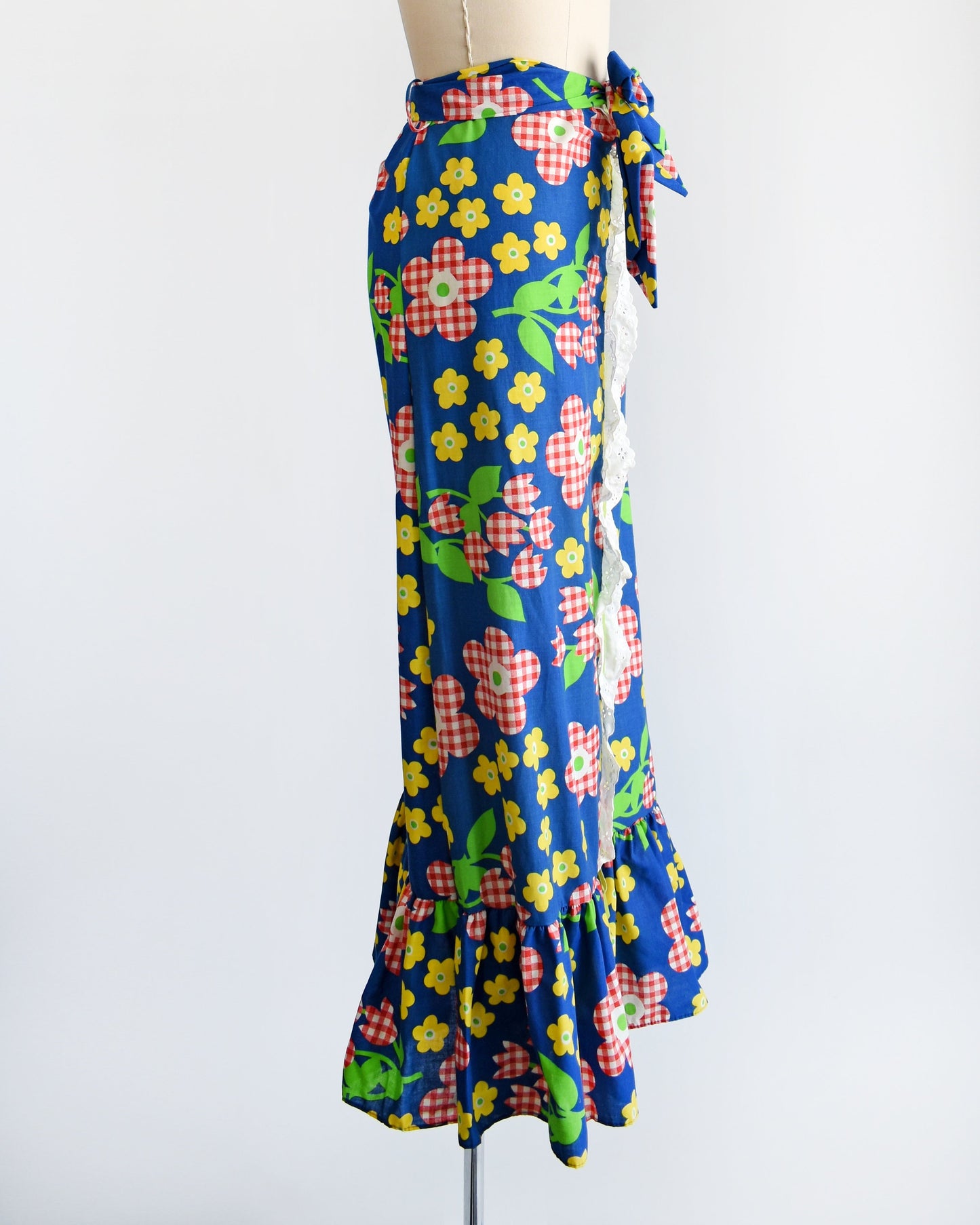Side view of a vintage 70s maxi skirt that is blue with a red and white gingham floral print, along with yellow and green flowers with green leaves and stems. White eyelet trim down the middle of the skirt with a velvet green ribbon down the middle.