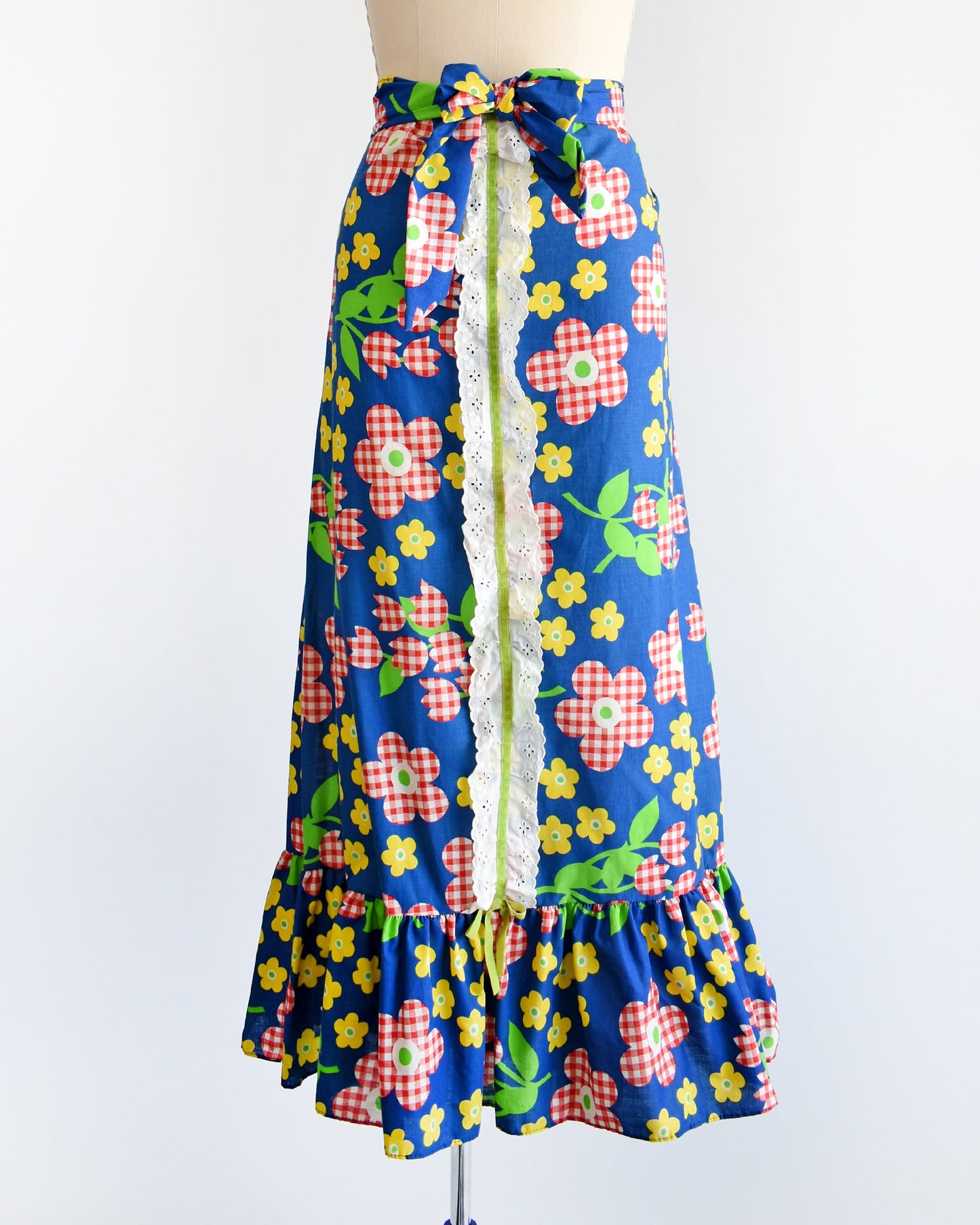 A vintage 70s maxi skirt that is blue with a red and white gingham floral print, along with yellow and green flowers with green leaves and stems. White eyelet trim down the middle of the skirt with a velvet green ribbon down the middle. Matching belt