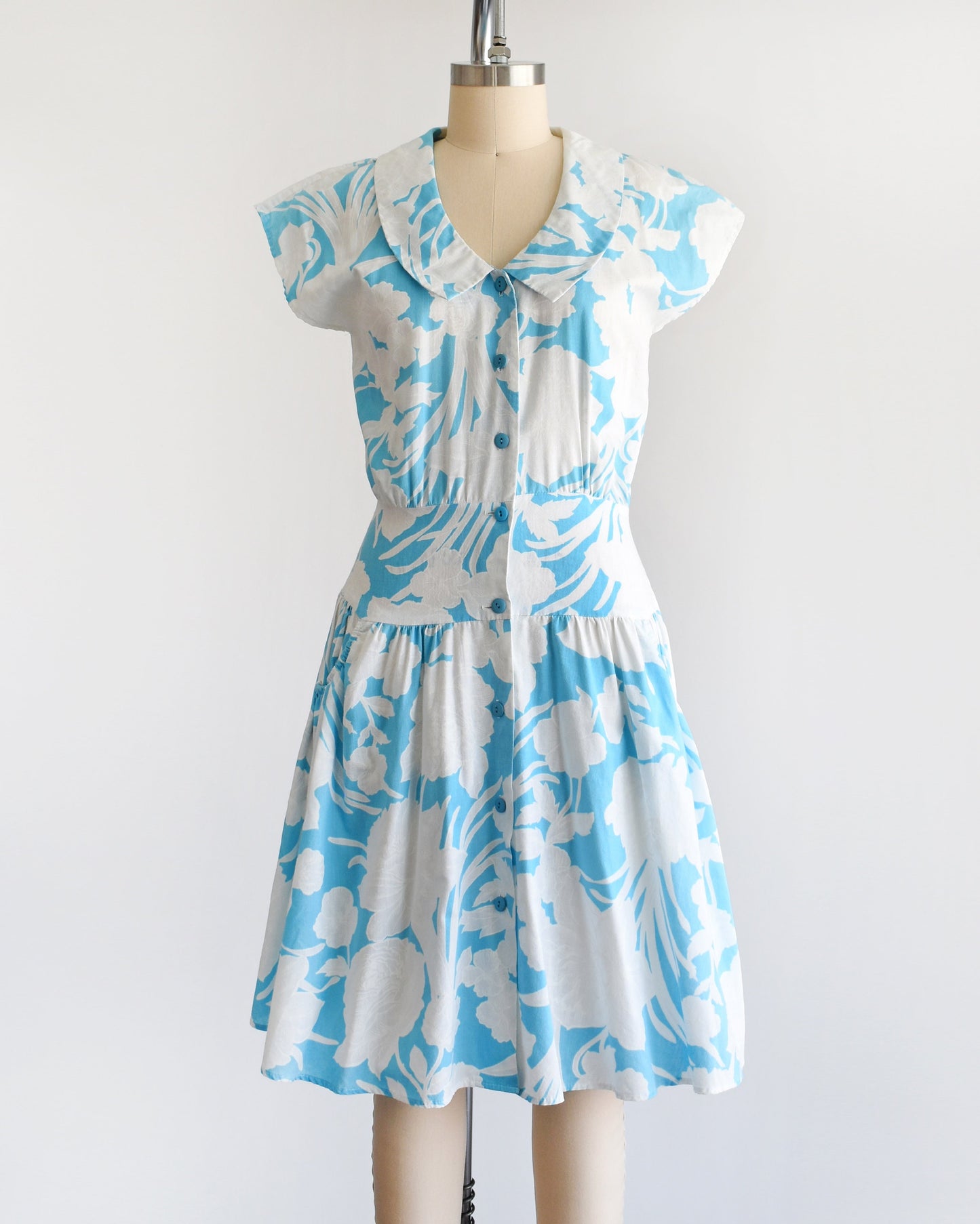 A vintage 80s floral dress that is light blue with white floral print. Peter Pan style collar with blue buttons down the front. Cap sleeves. Wide waistband and drop waist skirt.