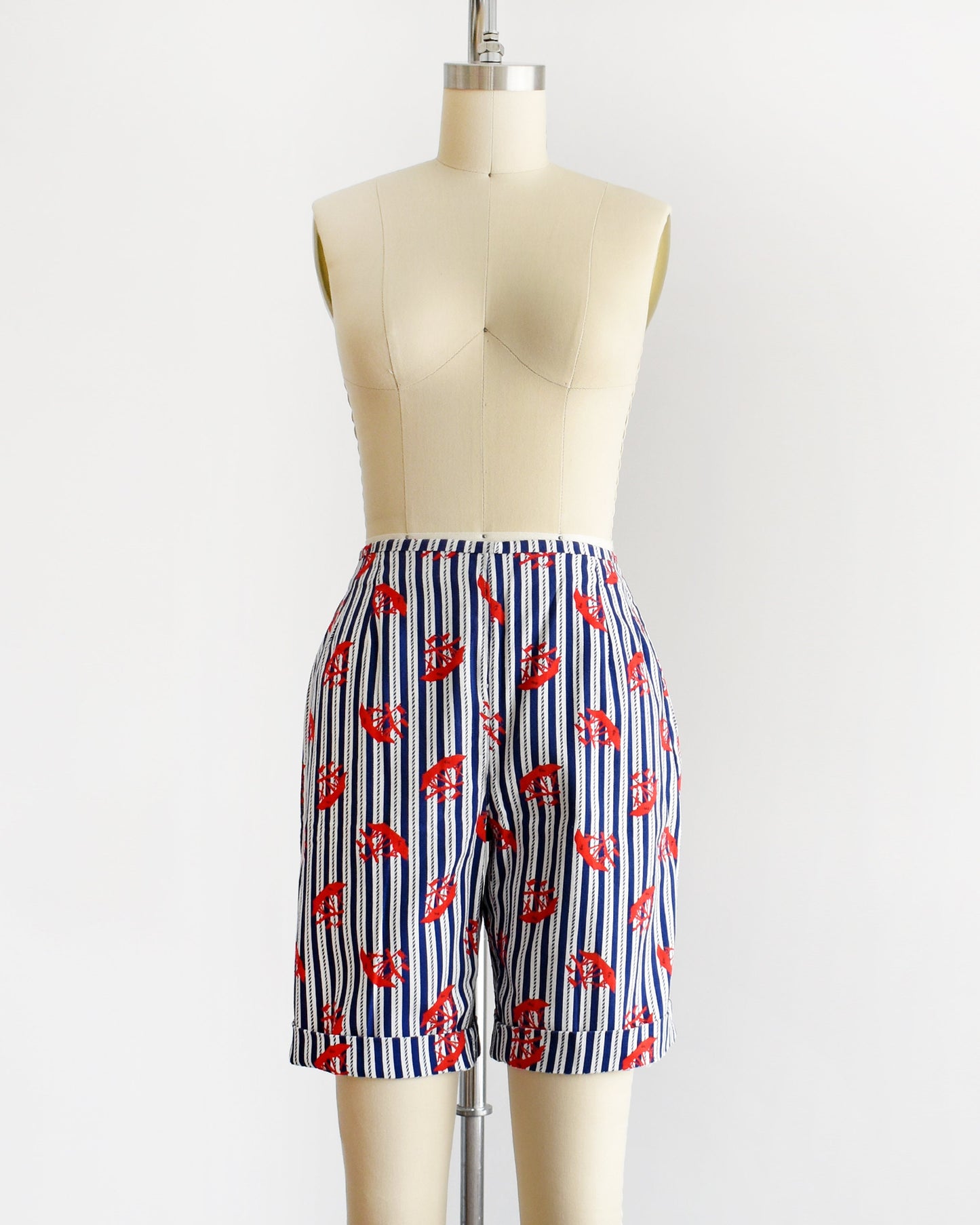 A pair of vintage 60s shorts that are navy blue and white vertical striped with a red clipper ship print. The shorts are modeled on a dress form. The shorts are cuffed in this photo