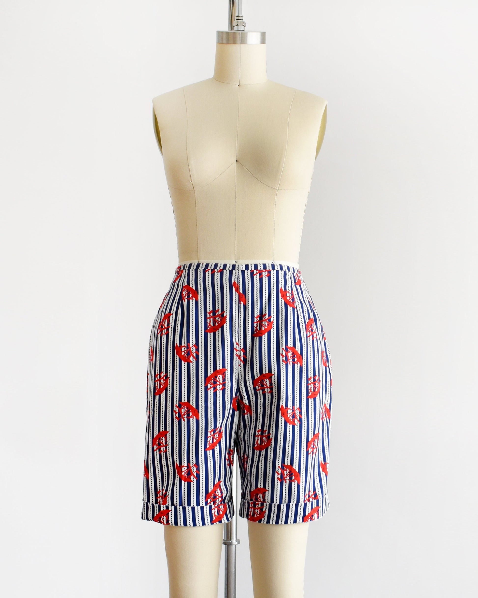 A pair of vintage 60s shorts that are navy blue and white vertical striped with a red clipper ship print. The shorts are modeled on a dress form. The shorts are cuffed in this photo