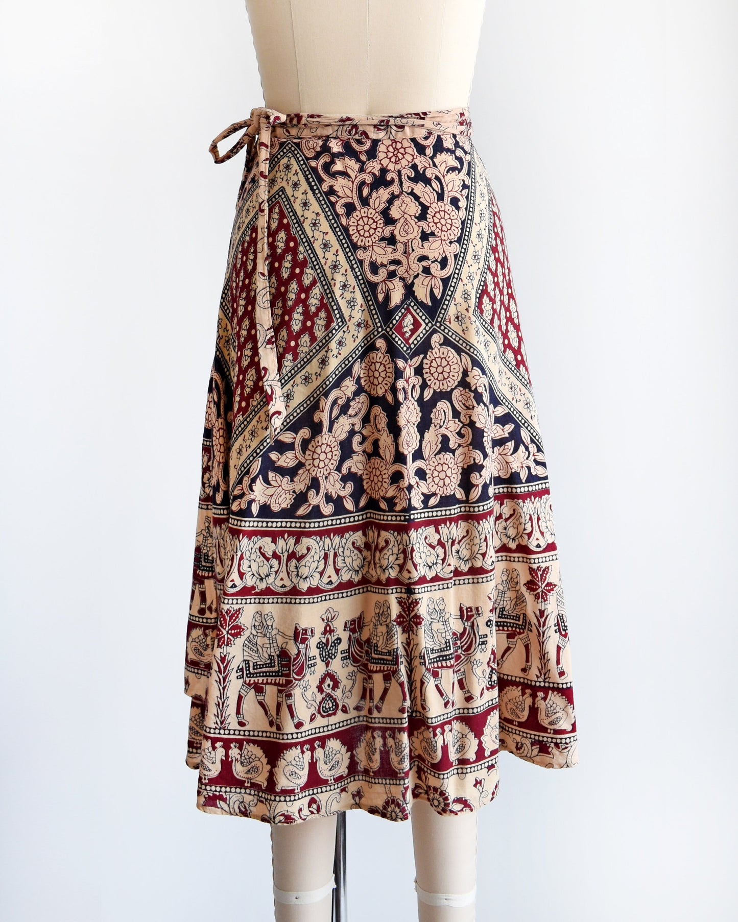 Back view of a vintage 1970s cotton wrap skirt that is beige with a large burgundy and black batik block floral print that covers the entire skirt, along border prints of people riding camels and a bird print. Skirt is on a dress form.