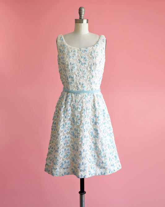 A vintage 60s white and blue floral lace dress that is sleeveless with a scoop neckline. Fitted waist with a matching double blue rope tie belt. Flared out skirt. Dress is modeled on a dress form