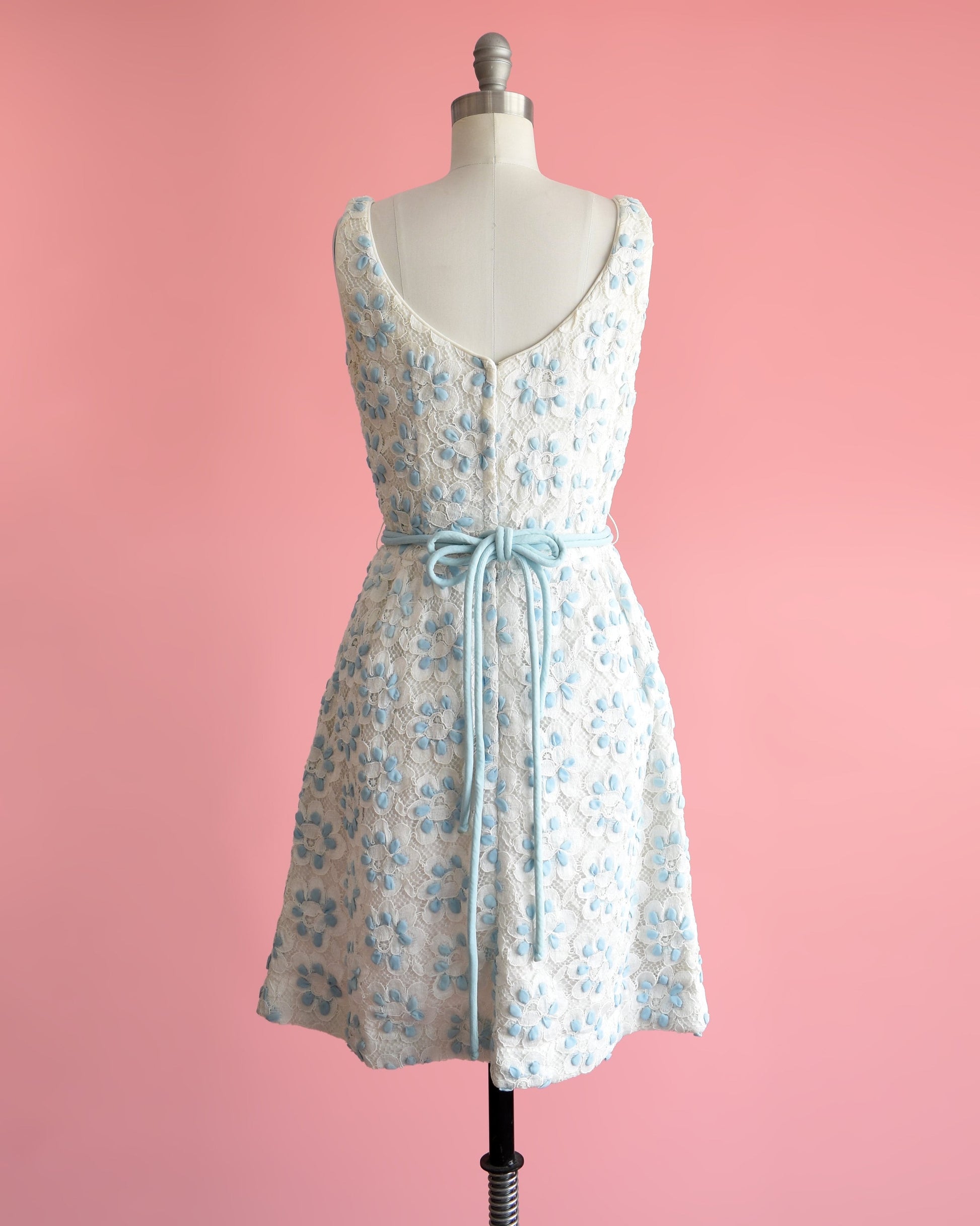 Back view of a vintage 60s white and blue floral lace dress that is sleeveless with a scoop neckline. Fitted waist with a matching double blue rope tie belt. Flared out skirt. Dress is modeled on a dress form