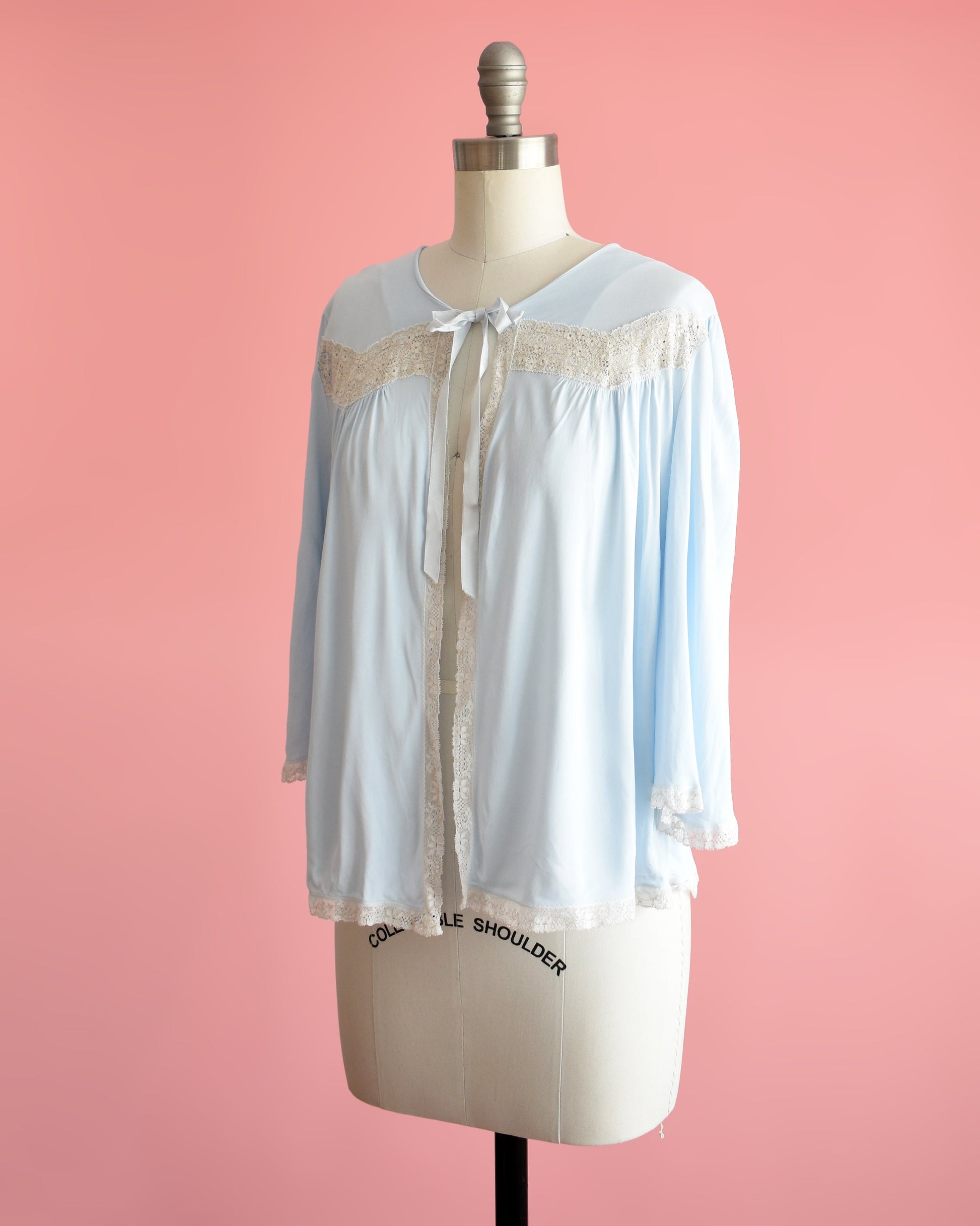 Side front view of a vintage 60s light blue bed jacket with cream lace trim around the front, down the front, around the hem, and cuffs. A ribbon tie is at the center of the collar. The garment is modeled on a dress form.