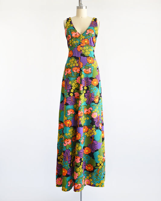A vintage 70s floral maxi dress that is black and has a vibrant flower power print in greens, blue, peach, orange, yellow, purple, and white. V-neckline. Dress is on dress form.