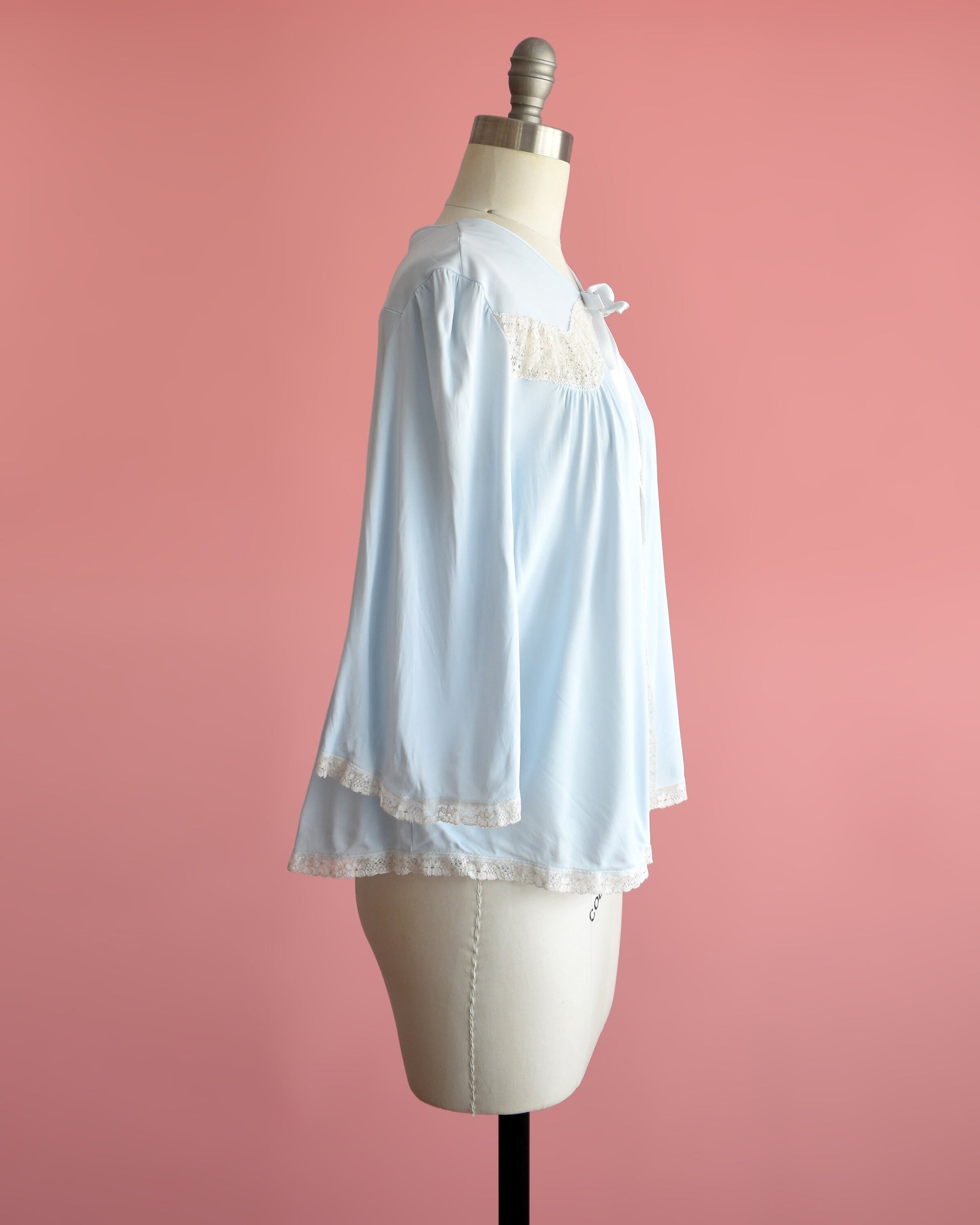 Side view of a vintage 60s light blue bed jacket with cream lace trim around the front, down the front, around the hem, and cuffs. A ribbon tie is at the center of the collar. The garment is modeled on a dress form.