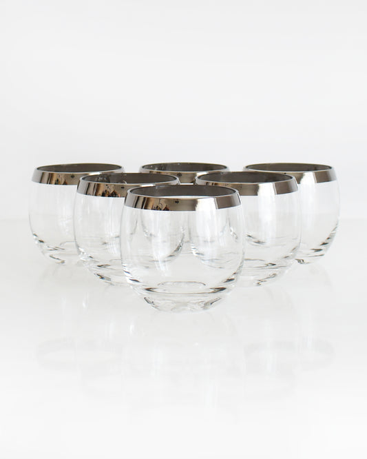 Six silver rim roly poly glasses that are round with flat bottoms on a white table against a white background