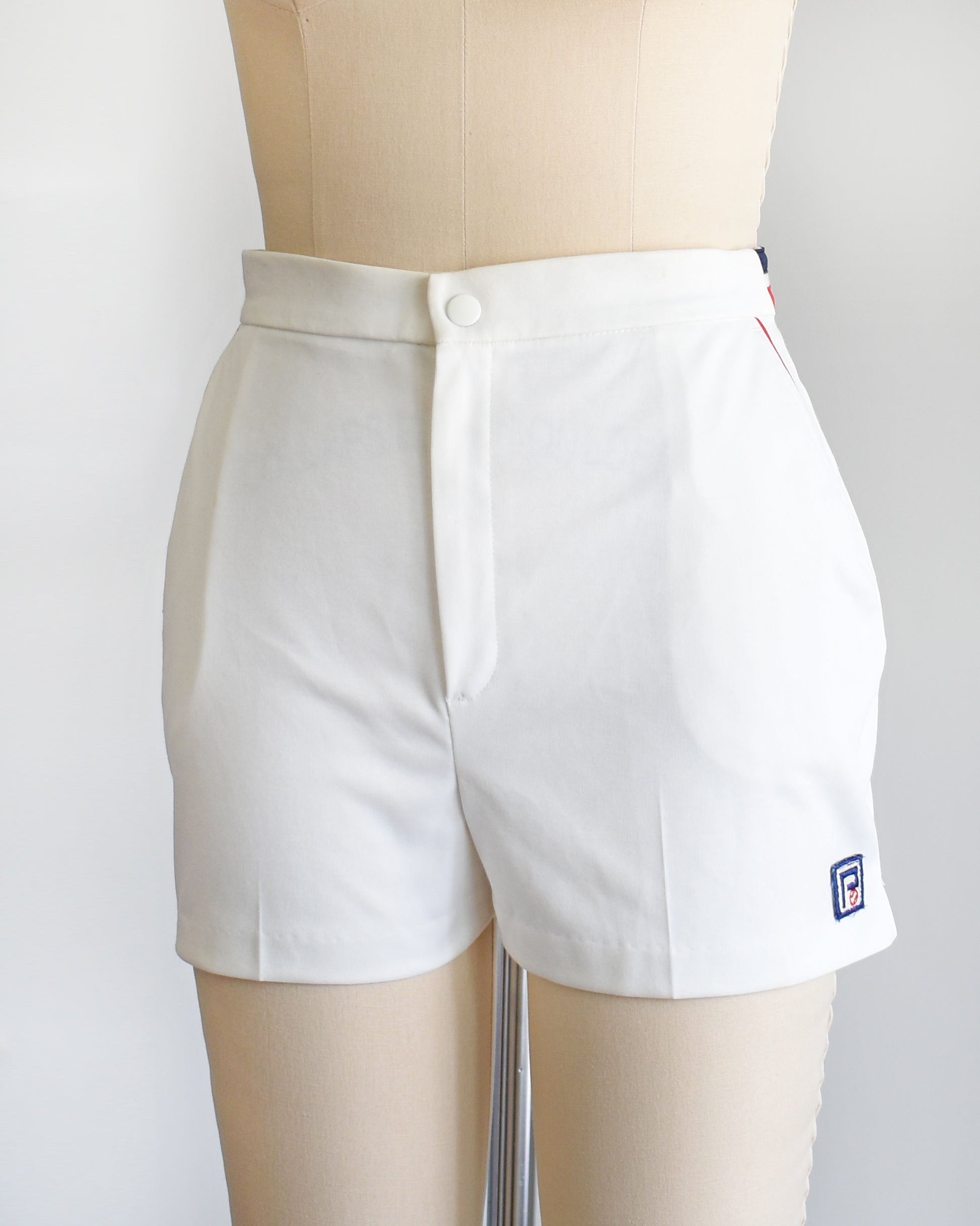 side front view of views of a vintage 1970s white tennis shorts on a dress form. The shorts have red and blue bands along the waist on the back and red trim around the pockets