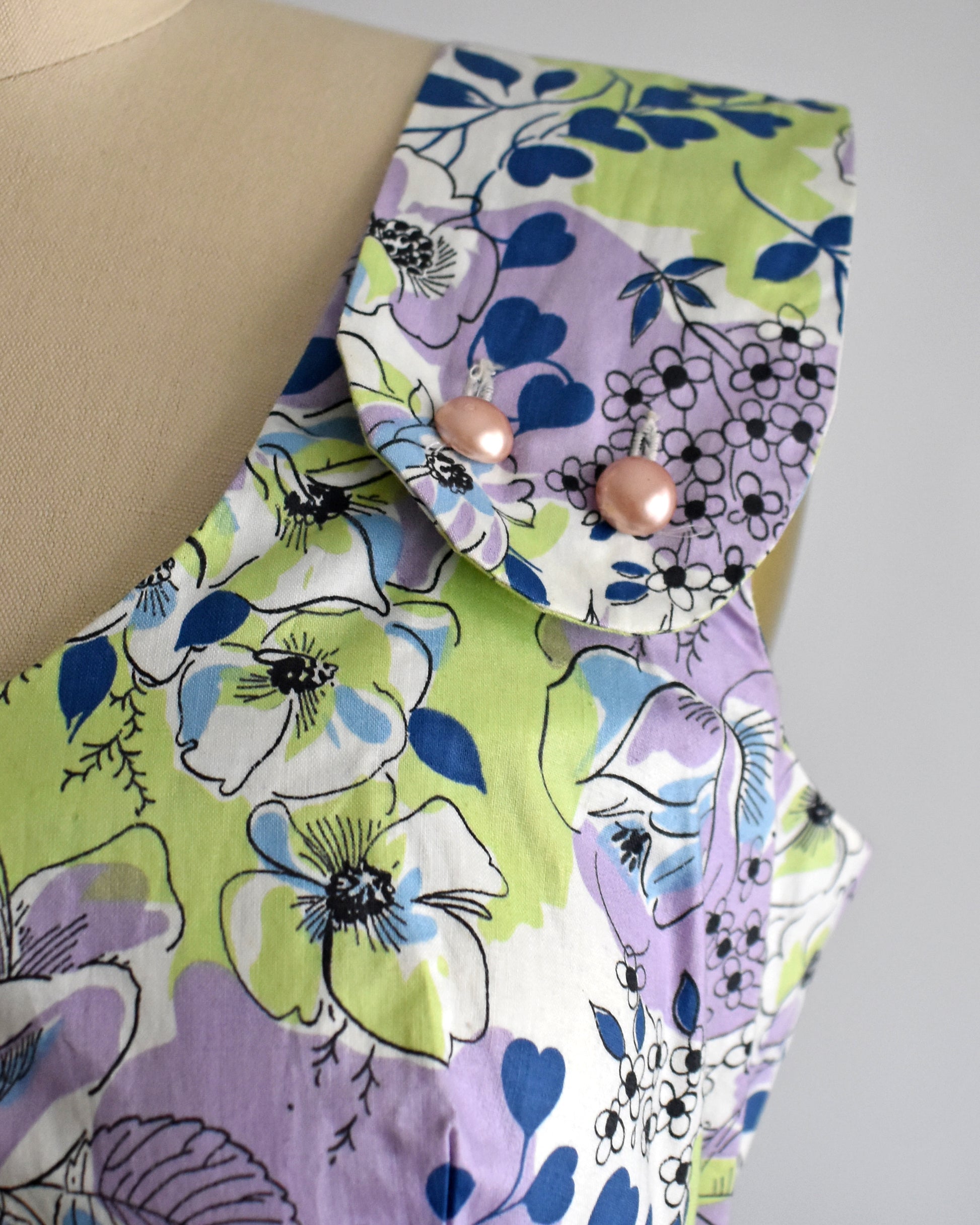 close up of the  pink strap buttons on a vintage 1950s purple, green, blue, and white floral cotton sundress on a dress form