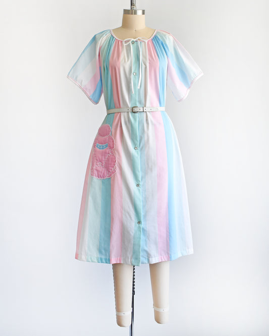 A vintage 1980s blue, pink, and white striped house dress with a pink poodle pocket on a dress form. There is a white belt around the waist which is not included.