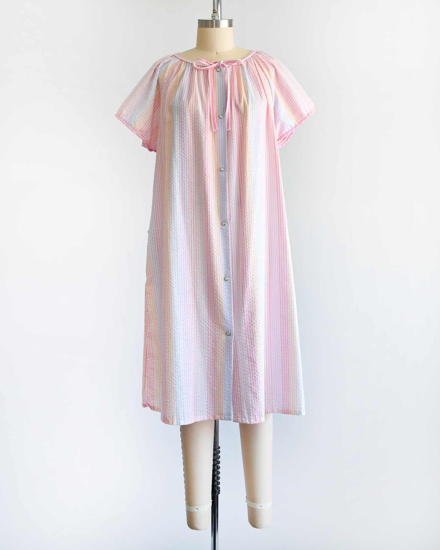 a vintage 1980s house dress that  features pastel rainbow stripes in blue, pink, orange, purple and white, along with a pink and white bow tie at the neck, and pearl snap buttons down the front. 